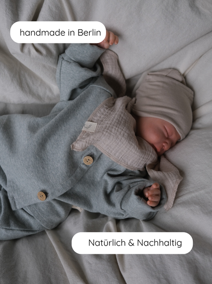 Neugeborenes in neutralem Bettzeug trägt Babyschlüttli Feinstrick - Mint Meliert mit Holzknöpfen, passender Mütze und Muslin-Bib. Labels: handmade in Berlin, Natürlich & Nachhaltig.
