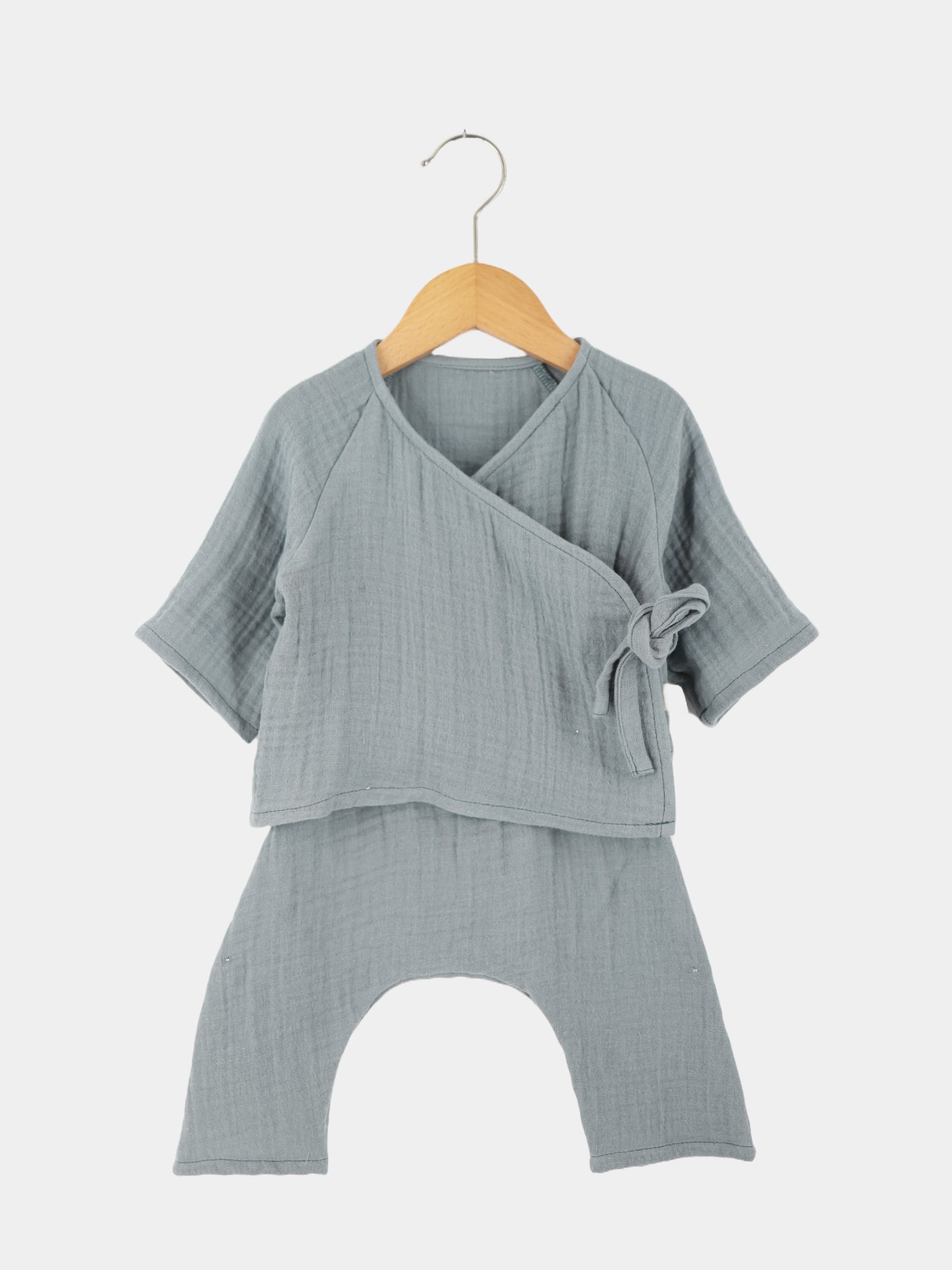 Babyset Baumwoll-Musselin - Eisblau: Detailaufnahme eines grauen Musselin-Wickel-Schlüttlis auf einem Holzkleiderbügel, perfekt für empfindliche Babyhaut, mit seitlicher Schleifenbindung und weichen, krempelbaren Ärmeln.