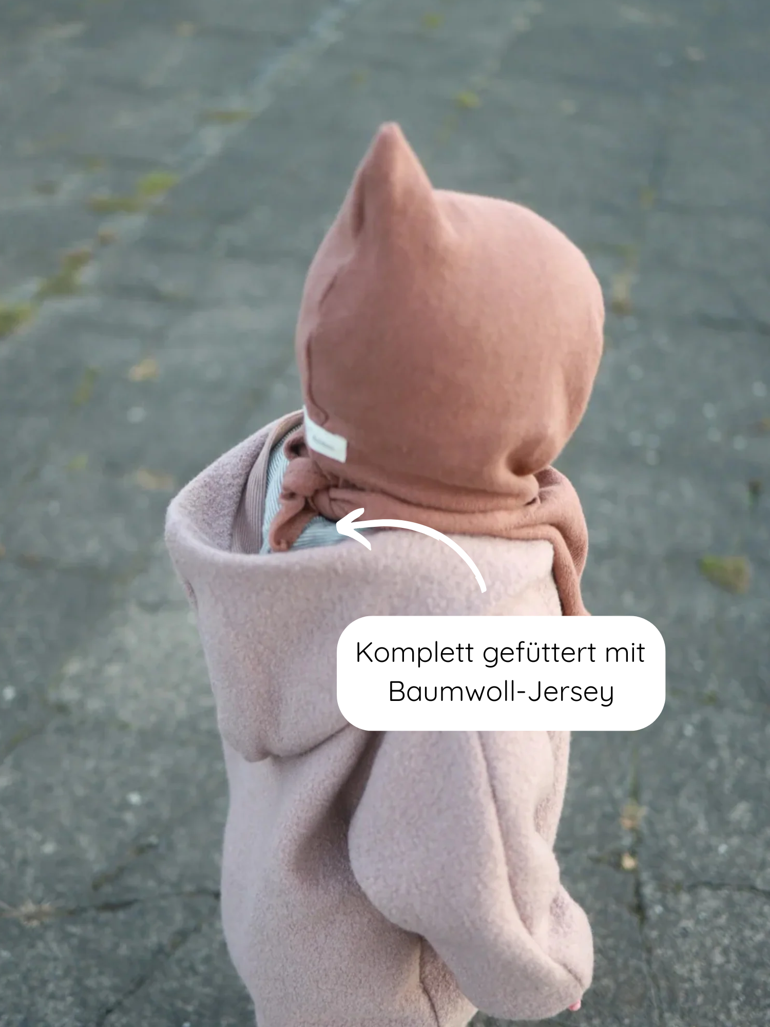 Kleinkind von hinten in Kuschelanzug Wollwalk - Puder aus Schurwolle mit Kapuze, gefüttert mit Baumwoll-Jersey, auf gepflastertem Untergrund. Weißer Pfeil zeigt auf das weiche Innenfutter.
