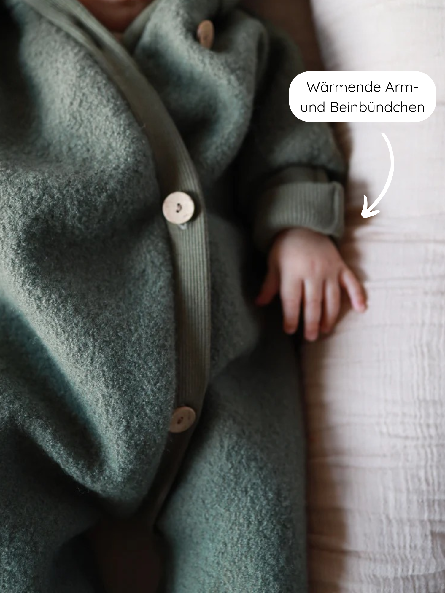 Kuschelanzug Wollwalk - Pistazie: Nahaufnahme eines Babys in einem waldgrünen, gefütterten Wollwalk-Overall mit Holzkopf-Knöpfen und wärmenden Arm- und Beinbündchen, der auf einer hellen Decke liegt.