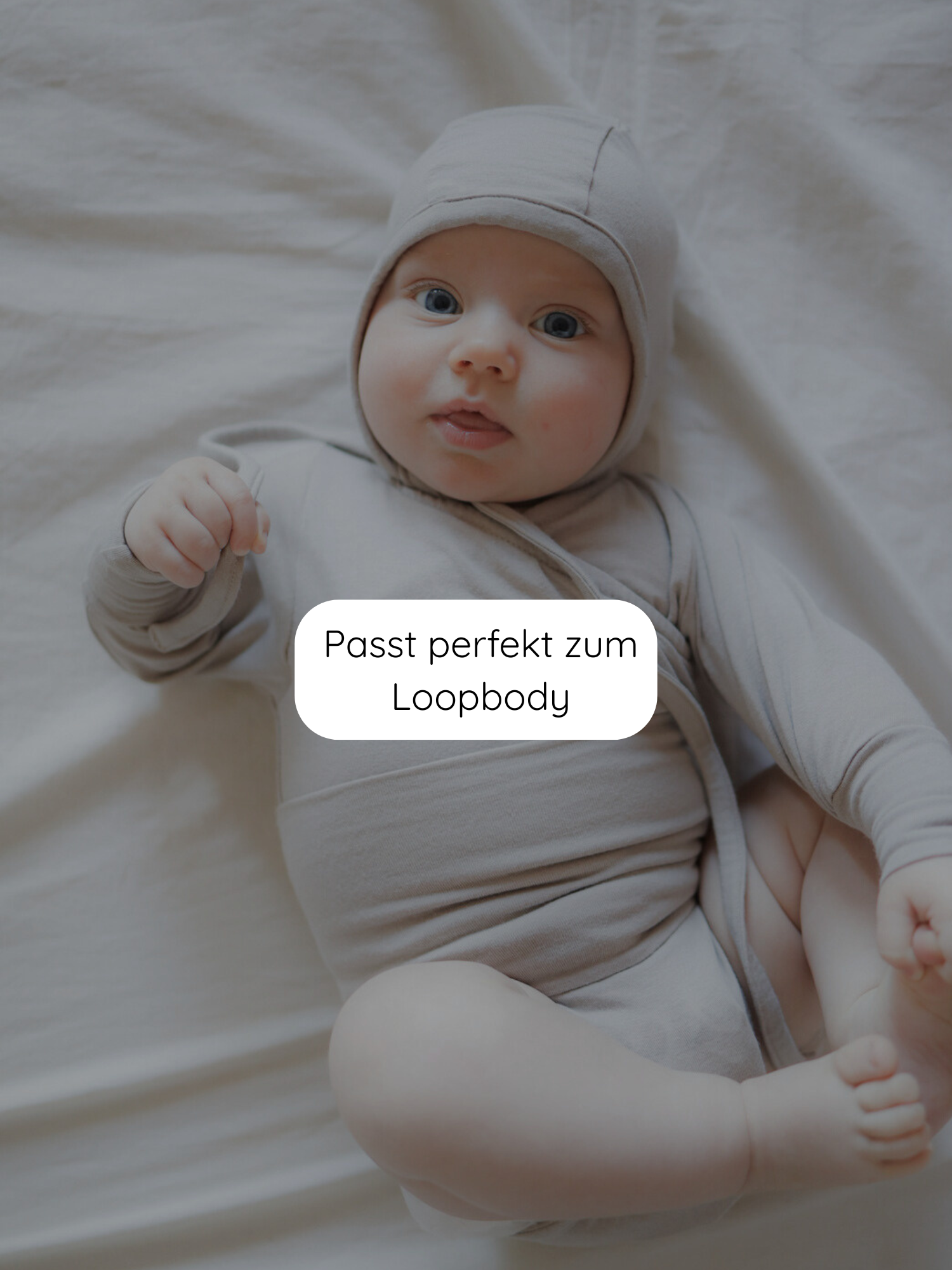 Babyhäubchen Kaschmir-Mix - Sand: Ein Baby liegt auf einem hellen Laken, trägt eine weiche, neutrale Mütze aus Kaschmir-Mix, passend zum Loopbody. Sanft beleuchtete, gemütliche Szene.