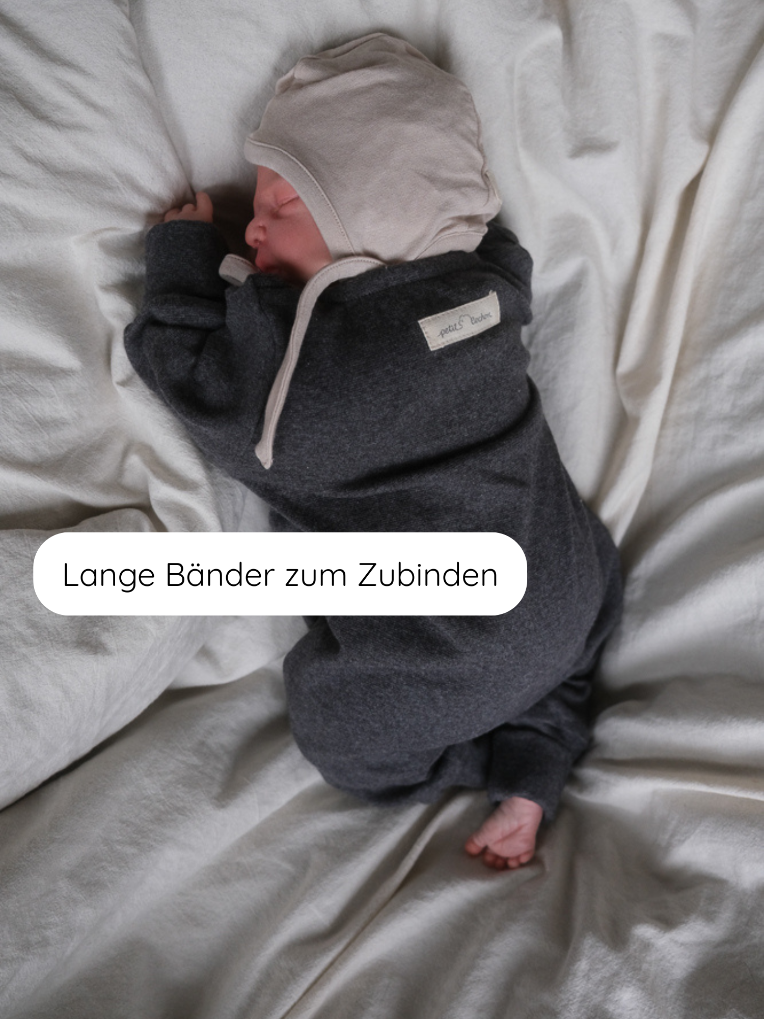 Schlafendes Baby auf weißem Laken, trägt ein Babyhäubchen Kaschmir-Mix - Sand mit langen Bändern und passendem dunkelgrauen Einteiler, illustriert durch Textblase „Lange Bänder zum Zubinden“.