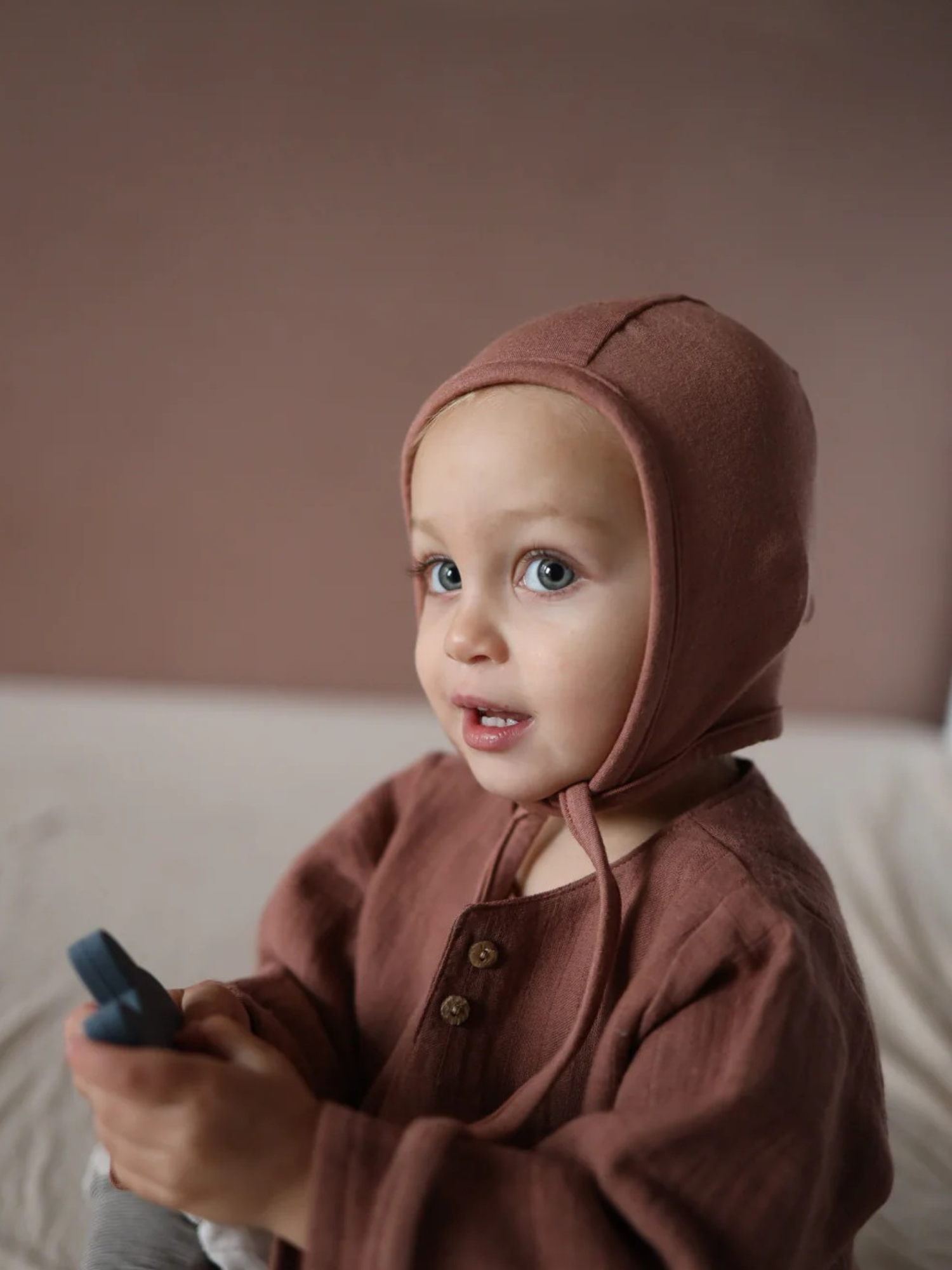 Kleinkind trägt ein Babyhäubchen Kaschmir-Mix - Zimt mit langen Bändern und weichem Stoff, passend zum Loopbody. Das Häubchen schmiegt sich sanft an den Kopf und bietet Wärme.