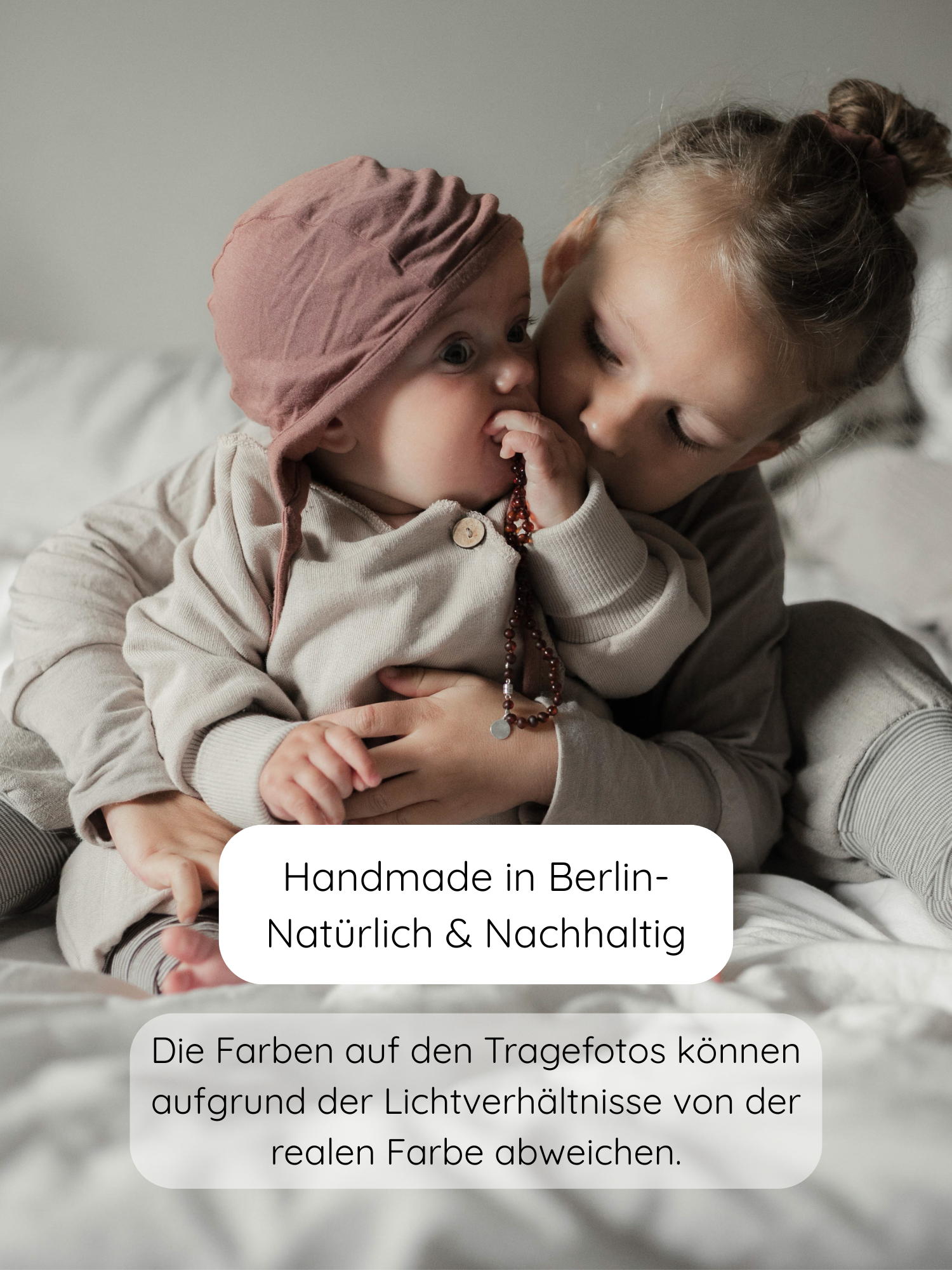 Zwei Kinder in kuscheligen Outfits, das Baby trägt das Babyhäubchen Kaschmir-Mix - Zimt mit einer rosafarbenen Haube. Der ältere Bruder hält das Baby sanft, während es Perlen festhält.