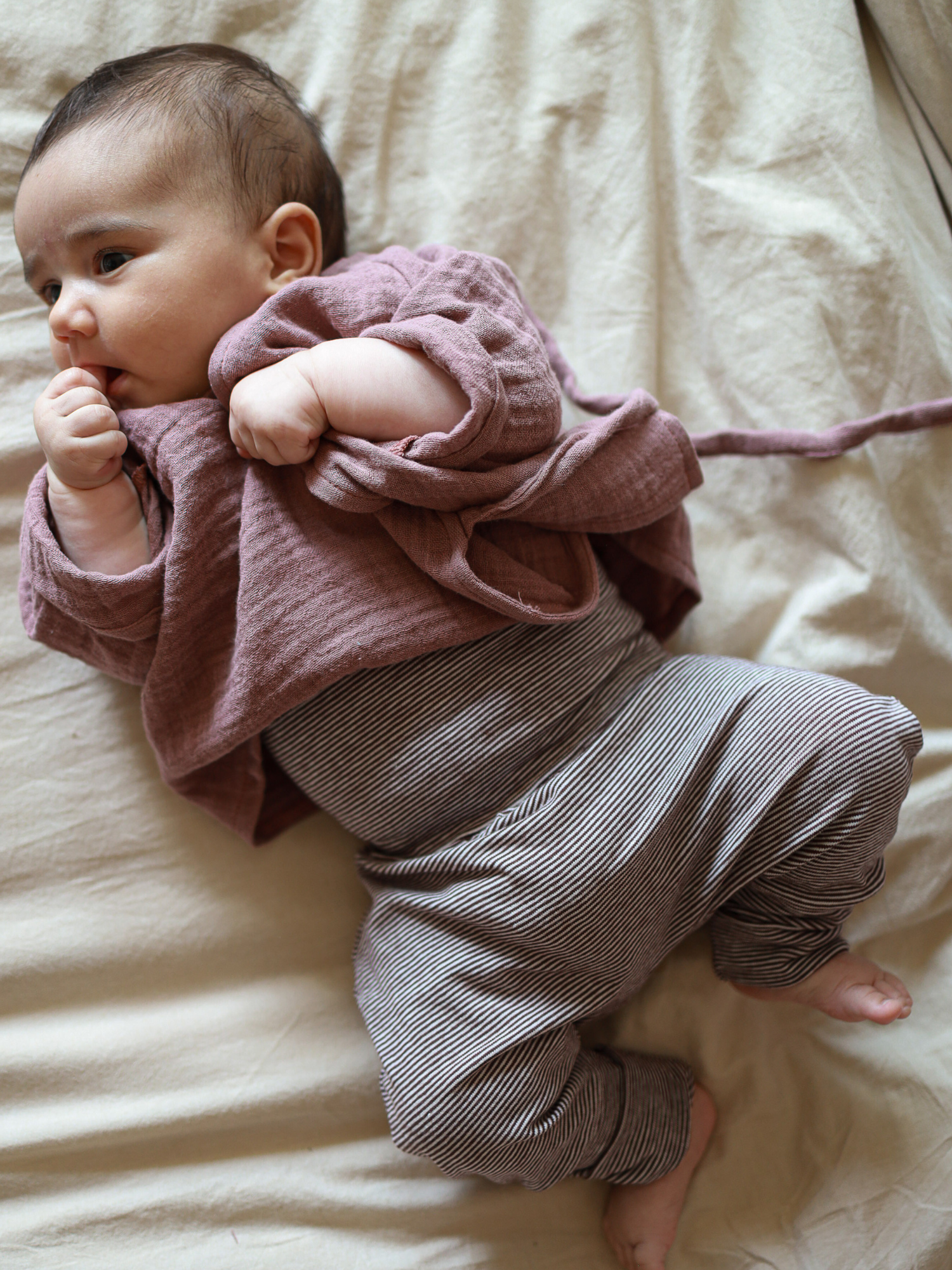 Baby trägt Babyknicker Bio-Baumwolle - Schoko Ringel, bequem auf heller Decke liegend. Die grauen und weißen Streifen sind sichtbar. Hand am Mund, Beine gemütlich hochgezogen.