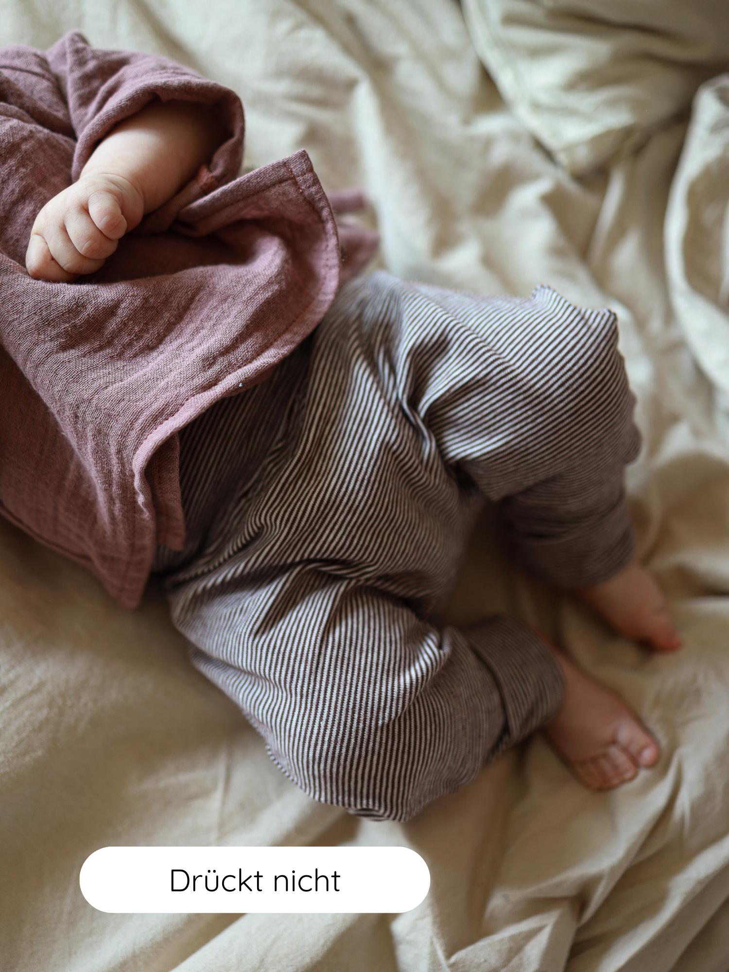 Baby liegt auf weicher Bettwäsche, trägt Babyknicker Bio-Baumwolle - Schoko Ringel. Die Hose ist grau-weiß gestreift, elastisch und bequem. Auf-Screen-Label: „Drückt nicht“. Perfekt für empfindliche Babyhaut.
