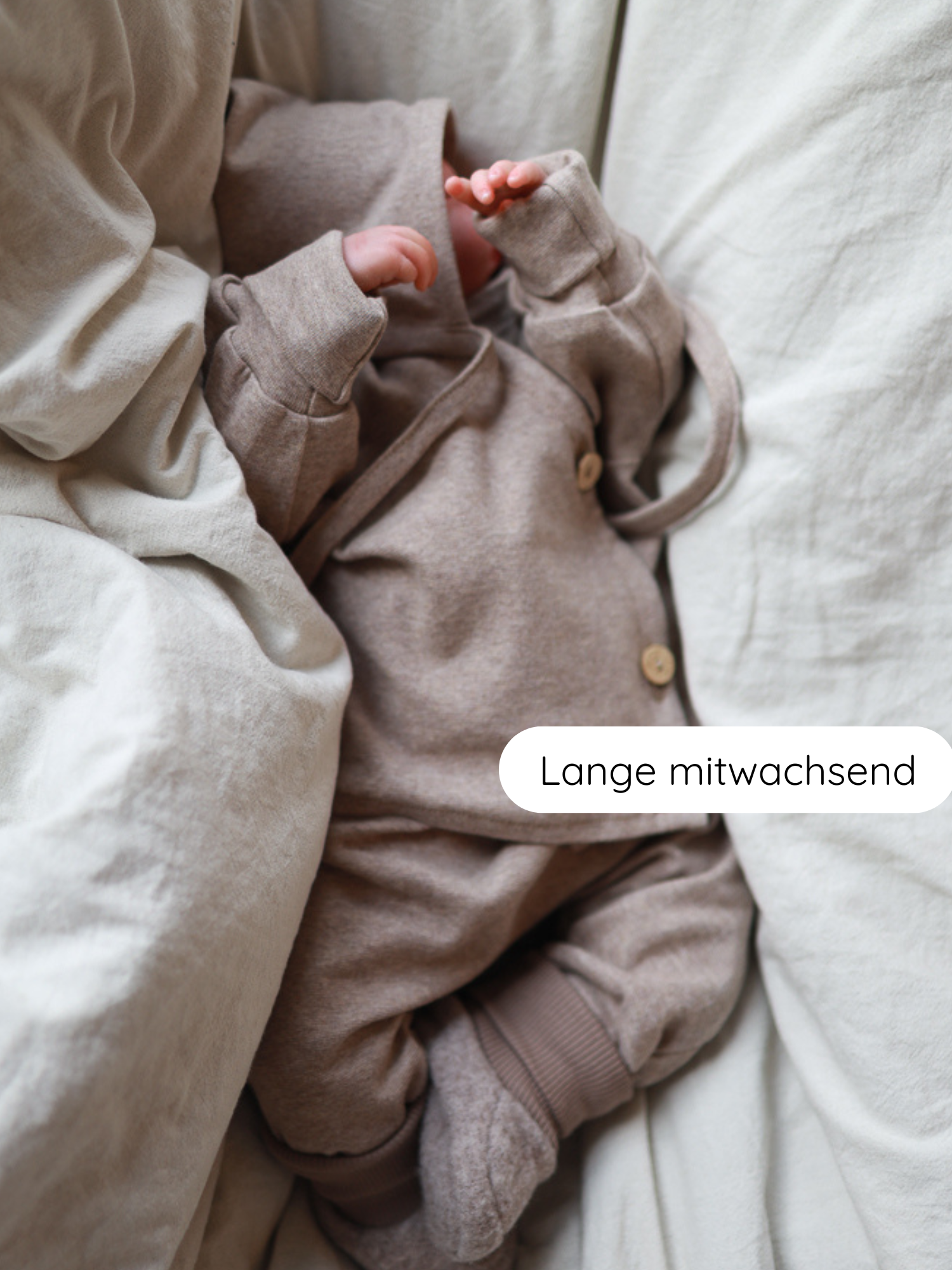 Neugeborenes in einem Babyknicker Feinstrick - Hafer Meliert Set mit Kapuzenjacke und passenden Hosen mit umklappbaren Bündchen, auf einer weißen Leinenoberfläche liegend.