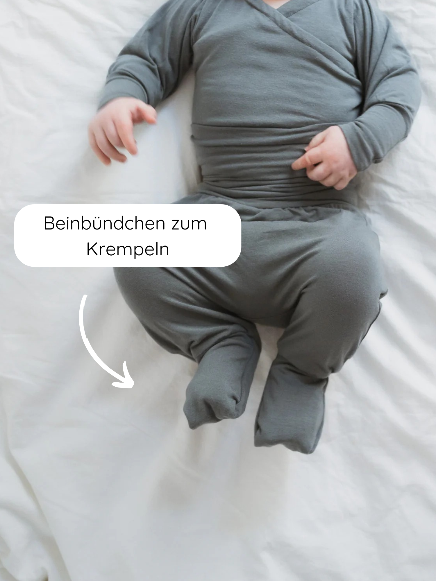 Baby in einem grauen, langärmeligen, fußbedeckten Babyknicker Kaschmir-Mix - Taube mit umklappbaren Beinbündchen, die sich hoch- oder runterrollen lassen, auf einem weißen Laken liegend.