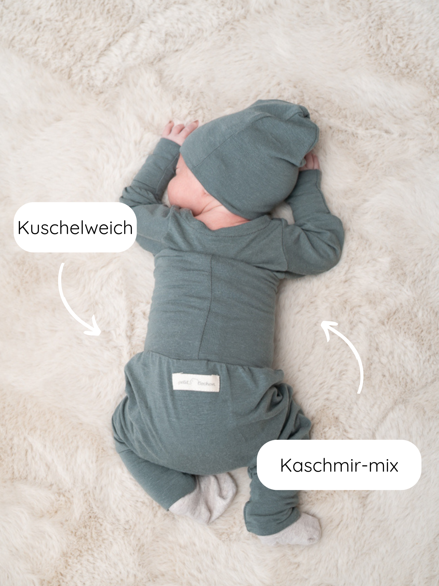 Neugeborenes auf cremefarbenem Kunstfell, gekleidet in Babyknicker Kaschmir-Mix - Taube, mit passendem Hut und Fußstrampler, betont durch Labels „Kuschelweich“ und „Kaschmir-mix“, die Weichheit und Luxus suggerieren.