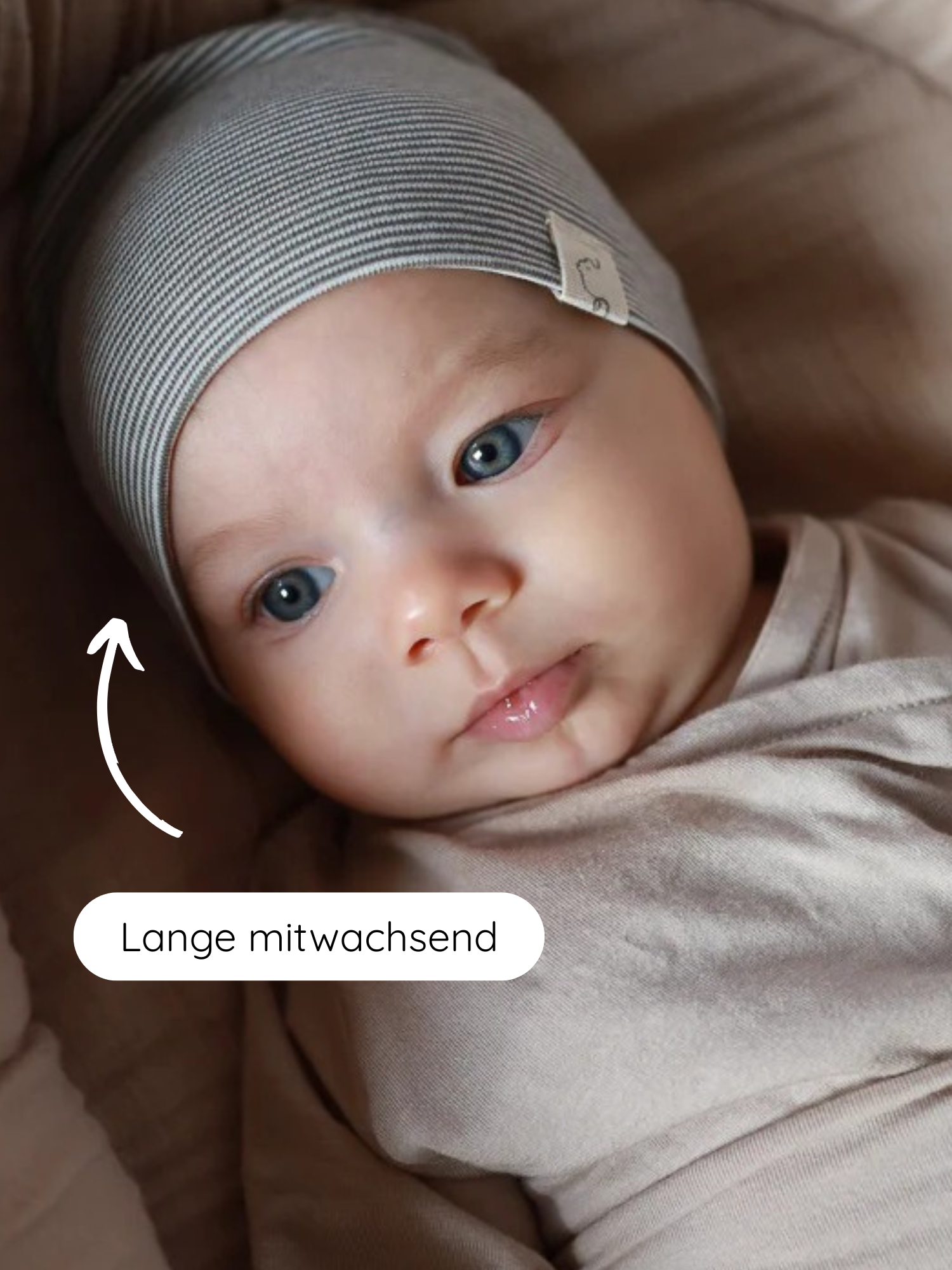 Baby mit blauen Augen, in beigem Wickel. Trägt Beanie Bio-Baumwolle - Ringel Maus, eine grauweiß gestreifte Mütze. Pfeil zeigt auf Mütze mit Aufschrift Lange mitwachsend, betont Langlebigkeit und Anpassungsfähigkeit.