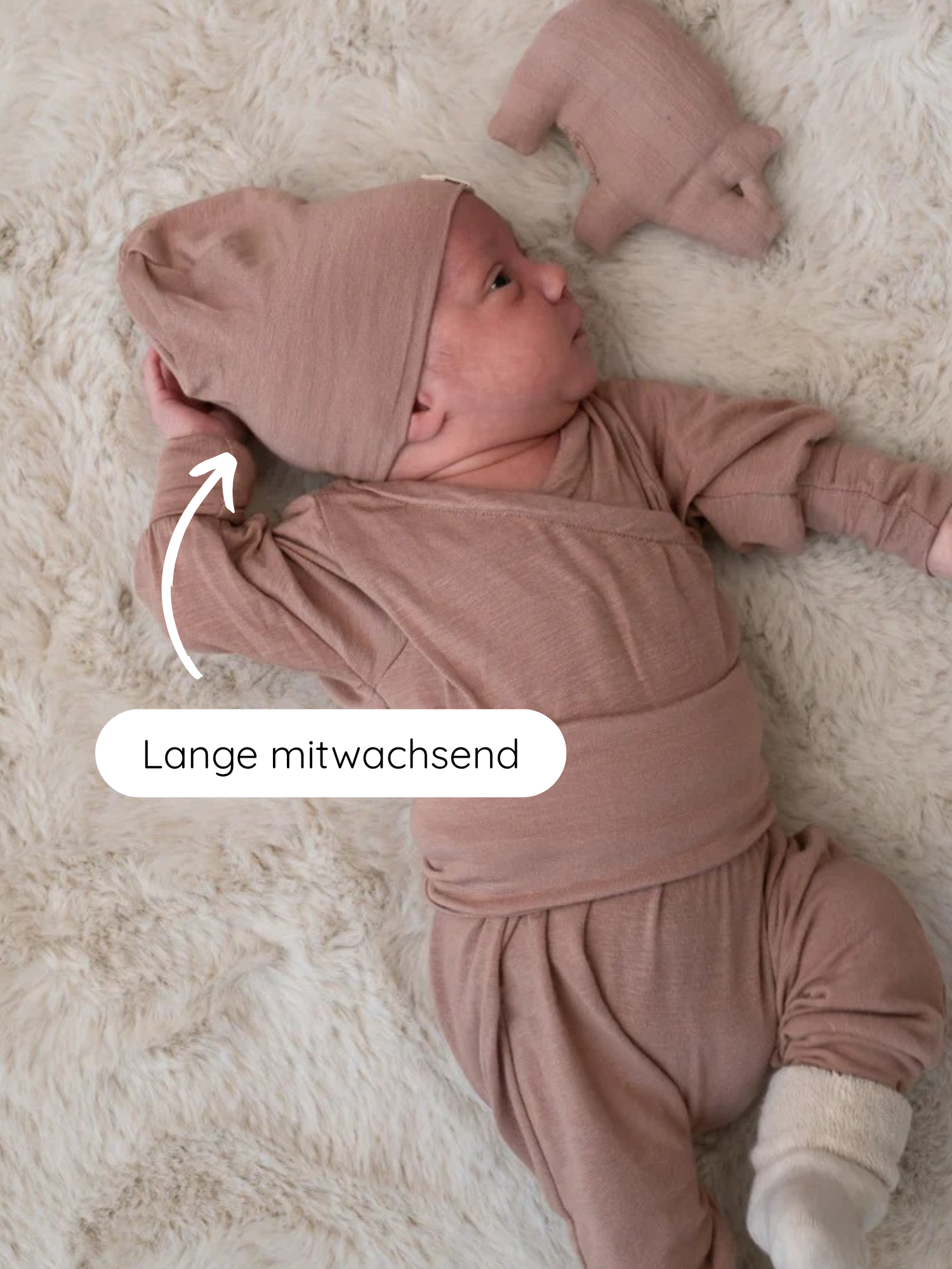 Neugeborenes auf cremefarbenem Kunstfell mit Beanie Kaschmir-Mix - Puder, langem mitwachsendem Hut und passendem Strickset. Stofftier-Schweinchen daneben. Lange mitwachsend zeigt auf den anpassbaren Hut.