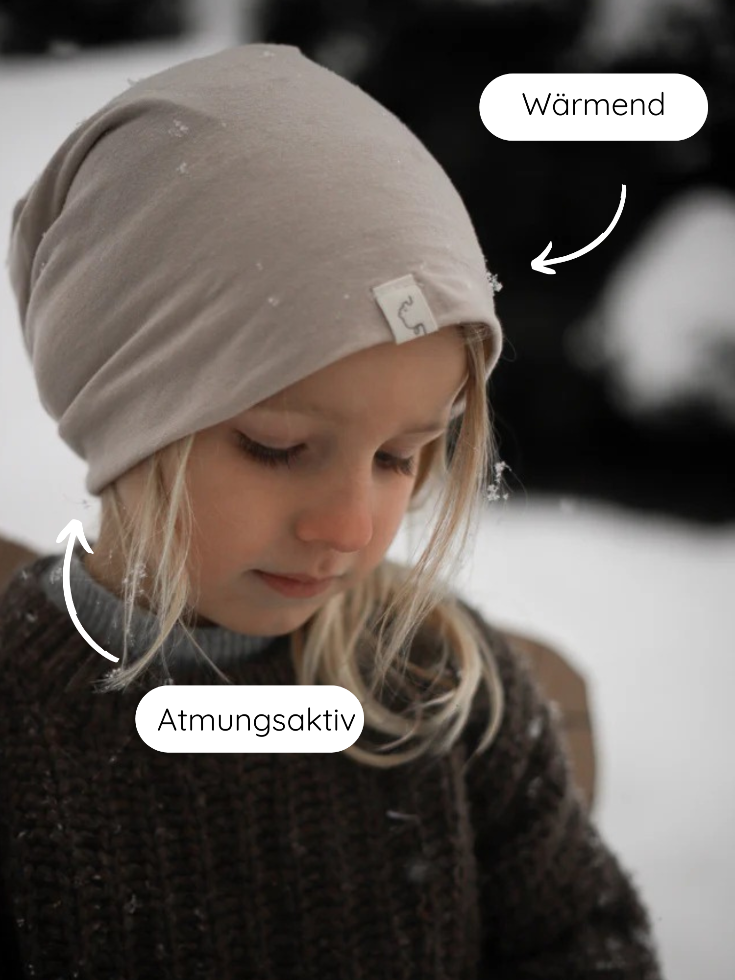 Kind im Schnee mit Beanie Kaschmir-Mix - Sand, eine taupefarbene Strickmütze. Grafische Hinweise heben die wärmenden und atmungsaktiven Eigenschaften hervor. Kombiniert mit einem dunklen, grob gestrickten Pullover.