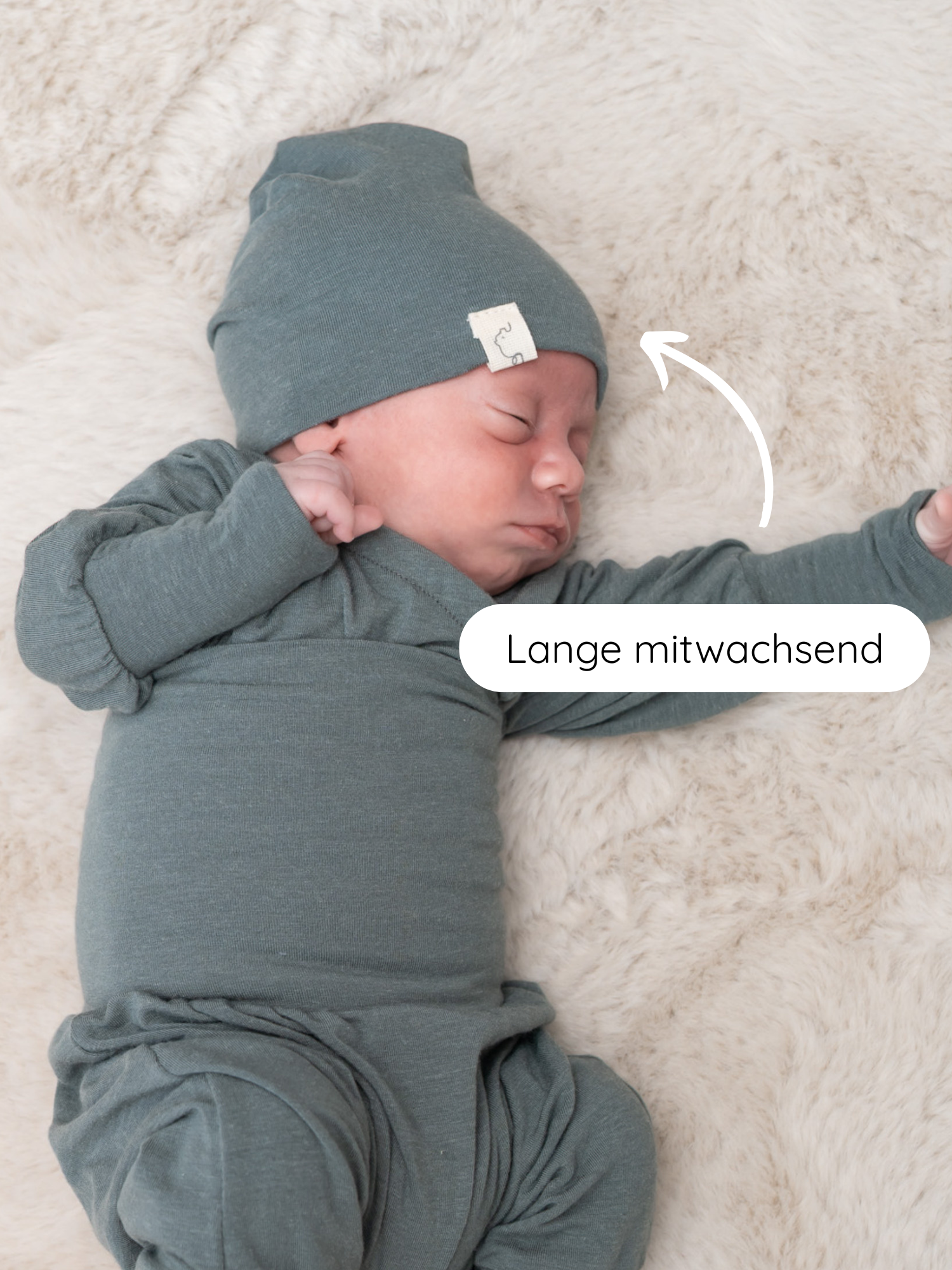 Schlafendes Neugeborenes in dehnbarem, salbeigrünem Grow-with-me-Outfit, das Beanie Kaschmir-Mix - Taube tragend. Ein Pfeil zeigt auf die Mütze, mit der Beschriftung Lange mitwachsend.