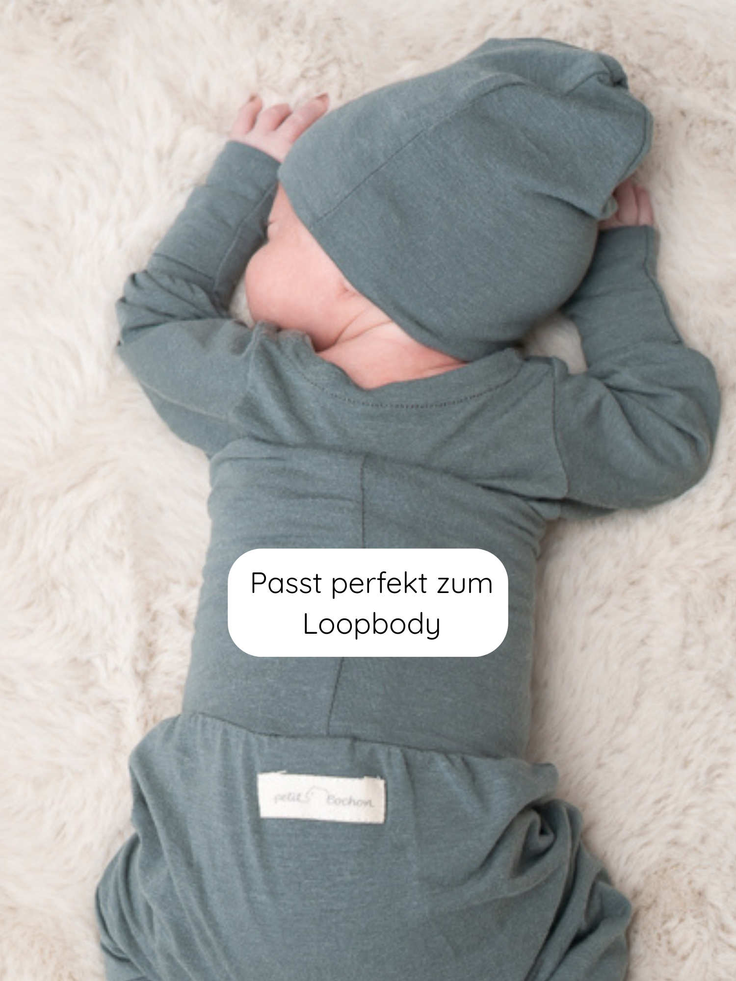 Baby liegt auf cremefarbenem Fellteppich, trägt Beanie Kaschmir-Mix - Taube. Weiche, dehnbare Mütze aus atmungsaktivem Material, handgefertigt in Berlin, passend zu Loopbody. Optimal für zarte Babyhaut.