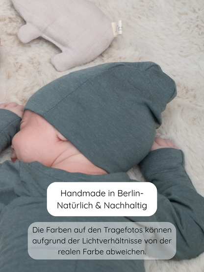 Baby auf cremefarbenem Teppich, trägt die Beanie Kaschmir-Mix - Taube in dezentem Grün, mit passendem Wickelanzug, neben einem beigen Kuscheltier. Text: Handmade in Berlin – Natürlich & Nachhaltig.