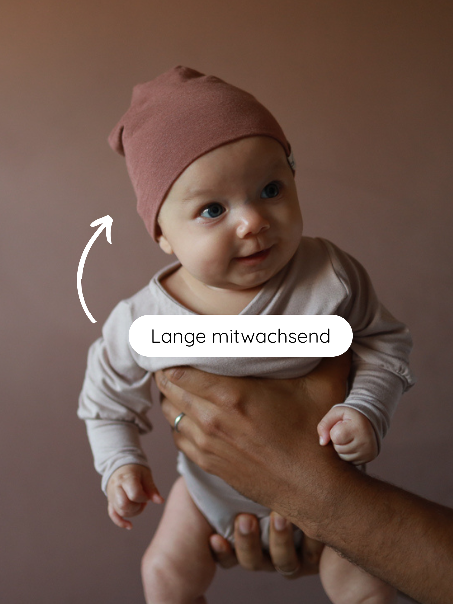 Kleines Baby trägt Beanie Kaschmir-Mix - Zimt, gehalten von Erwachsenenhand. Weiße Zeichnung zeigt auf die Mütze mit dem Text „Lange mitwachsend“, betont die dehnbare, komfortable Passform der Mütze.