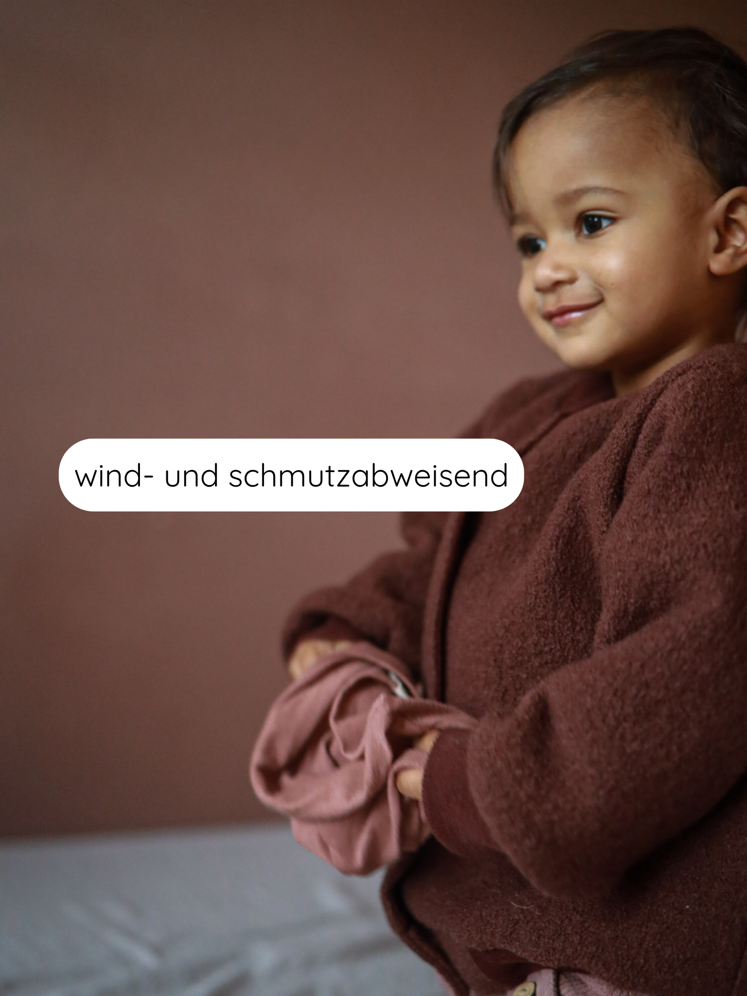 Kleines Kind trägt die Bomberjacke Wollwalk - Schoko, eine teddyfleeceartige Jacke. Es hält ein rosa Kleidungsstück. Die Jacke ist wind- und schmutzabweisend, ideal für aktive Kinder.