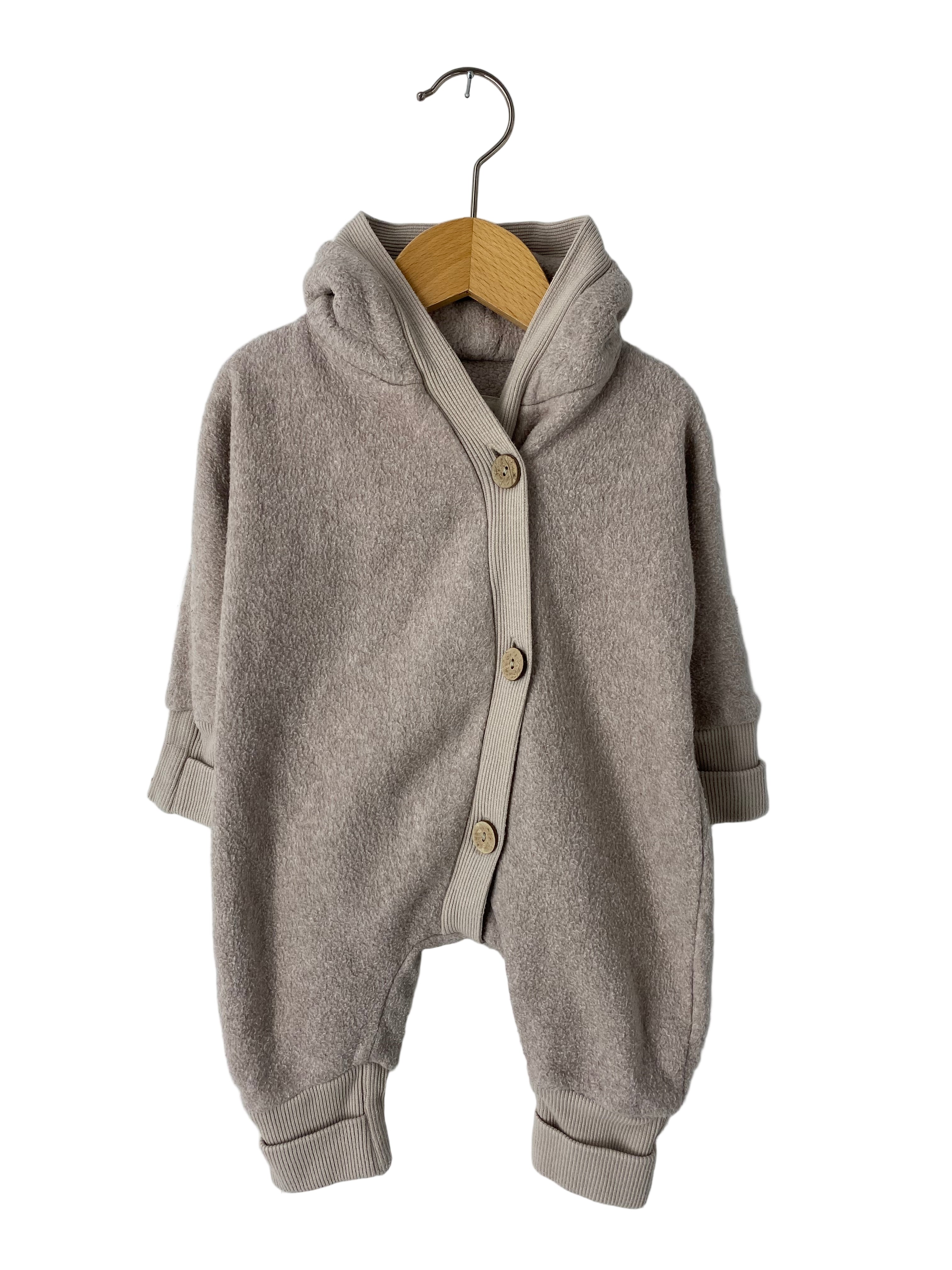 Kuschelanzug Bio-Plüsch Sand Gr 0M: Ein einteiliger Babystrampler aus weichem Frotteestoff in Beige, mit Kapuze, langen Ärmeln, Bündchen und schräger Knopfleiste mit drei Holzknöpfen, auf einem Holzkleiderbügel hängend.