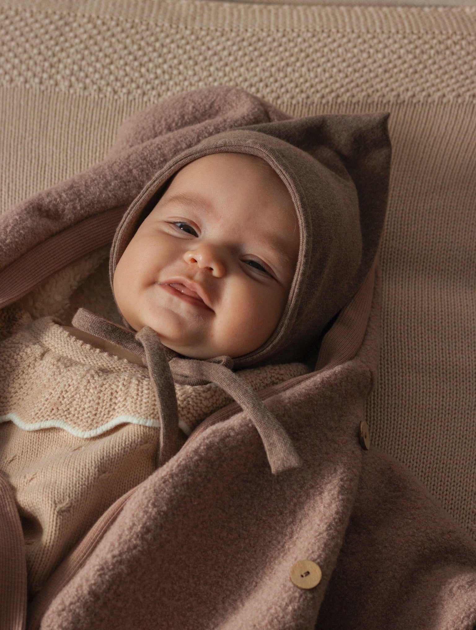 Lächelndes Baby in Zwergenmütze Feinstrick - Praline Meliert, mit charakteristischer Zipfelform und Kinnbändern, kombiniert mit passendem Wollmantel, ruhend auf strukturiertem, beigem Hintergrund für einen gemütlichen Look.