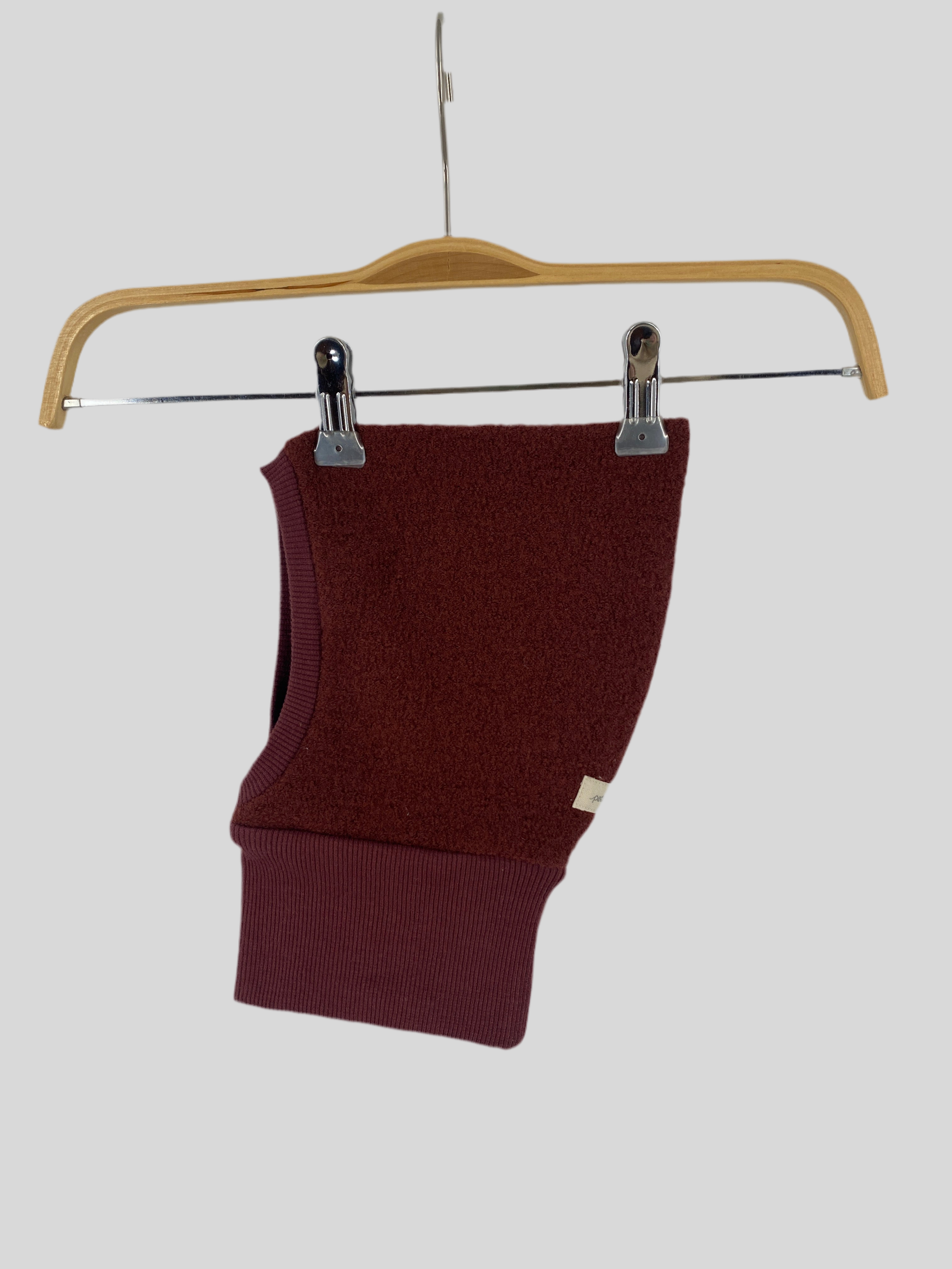 Schlupfmütze Wollwalk Burgund Gr 0M-6M auf einem Kleiderbügel, Detailansicht der gleichmäßigen Stoffstruktur und Verarbeitung, präsentiert auf einem Holzschwinger mit Metallgriff. Zustand: wie neu.