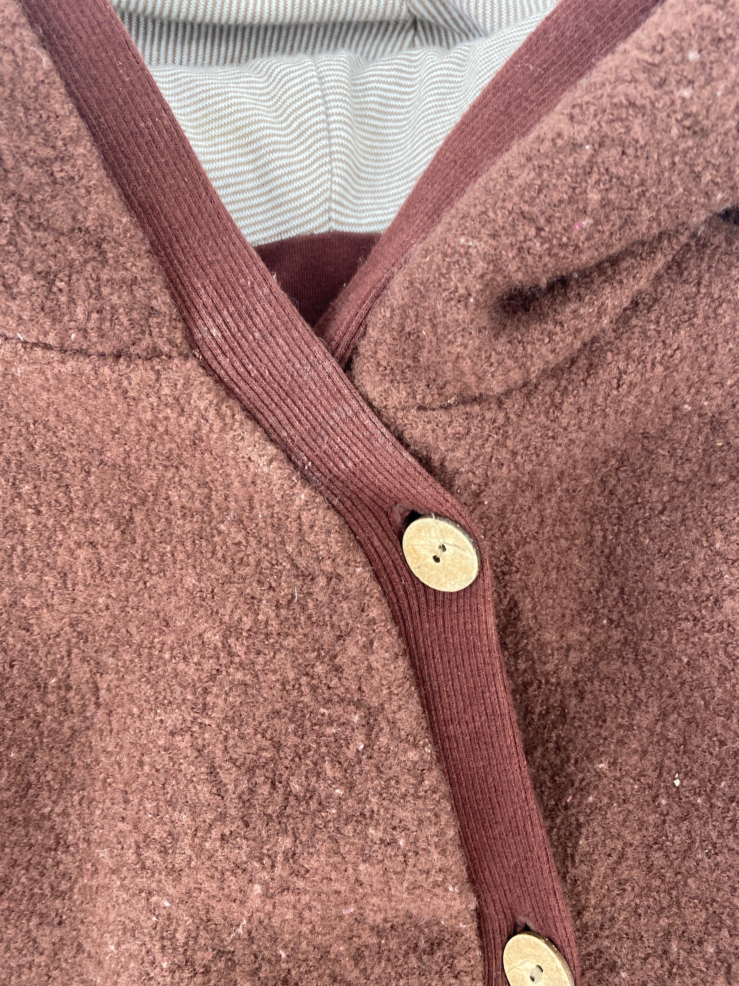 Nahaufnahme des (Kopie) Kuschelanzug Wollwalk Schoko Gr 1Y: brauner Bouclé-Cardigan mit geripptem Besatz, zwei Holzknöpfen und sichtbarem gestreiftem Innenfutter. Zustand: gut, Hals-Bündchen etwas fleckig.