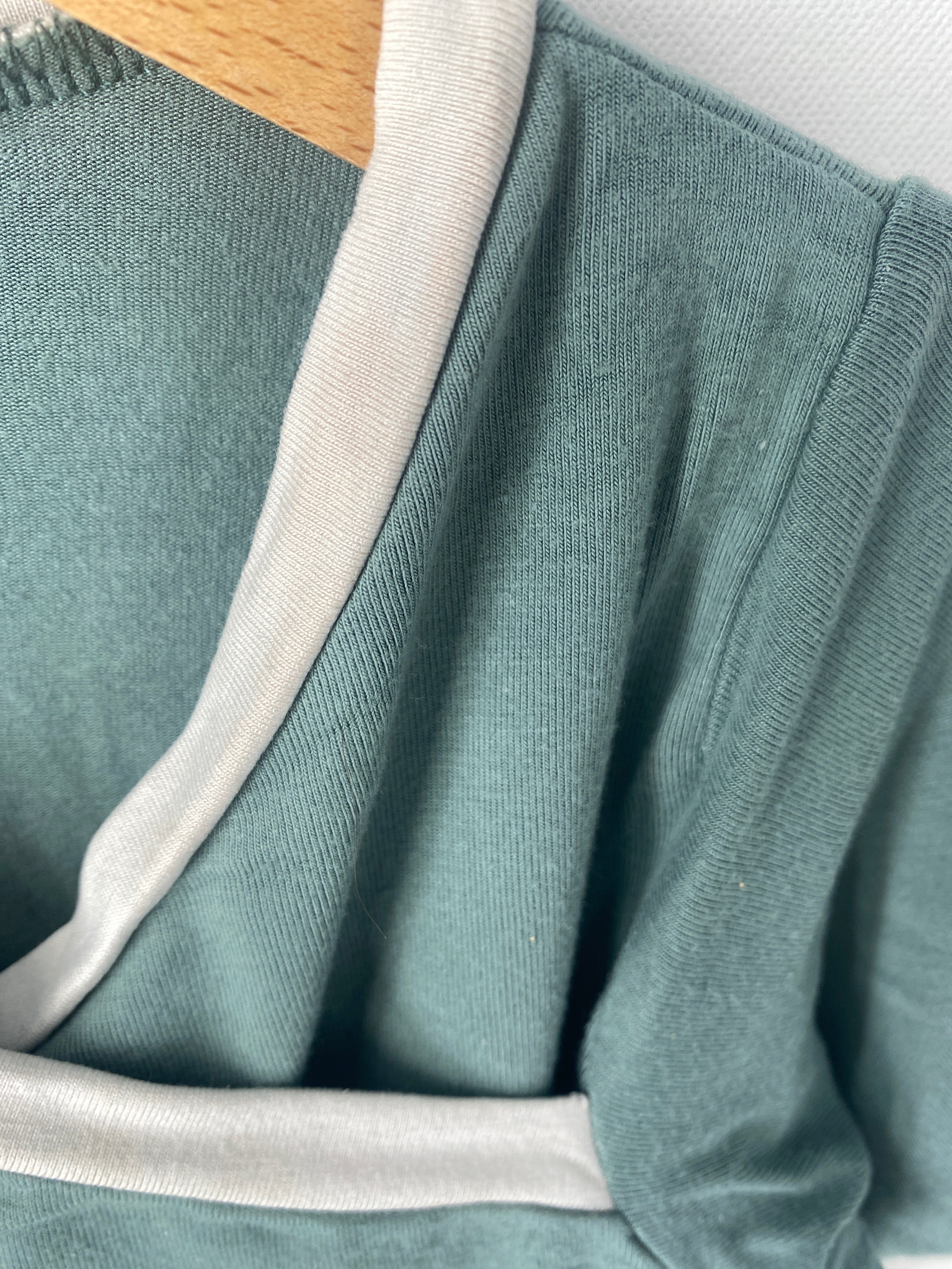 Nahaufnahme des Ringer Shirts Bambusviskose Jade Gr 3Y mit weißem Kontrastbesatz, leichte Flecken sichtbar, auf einem Holzbügel drapiert. Zustand: gut.