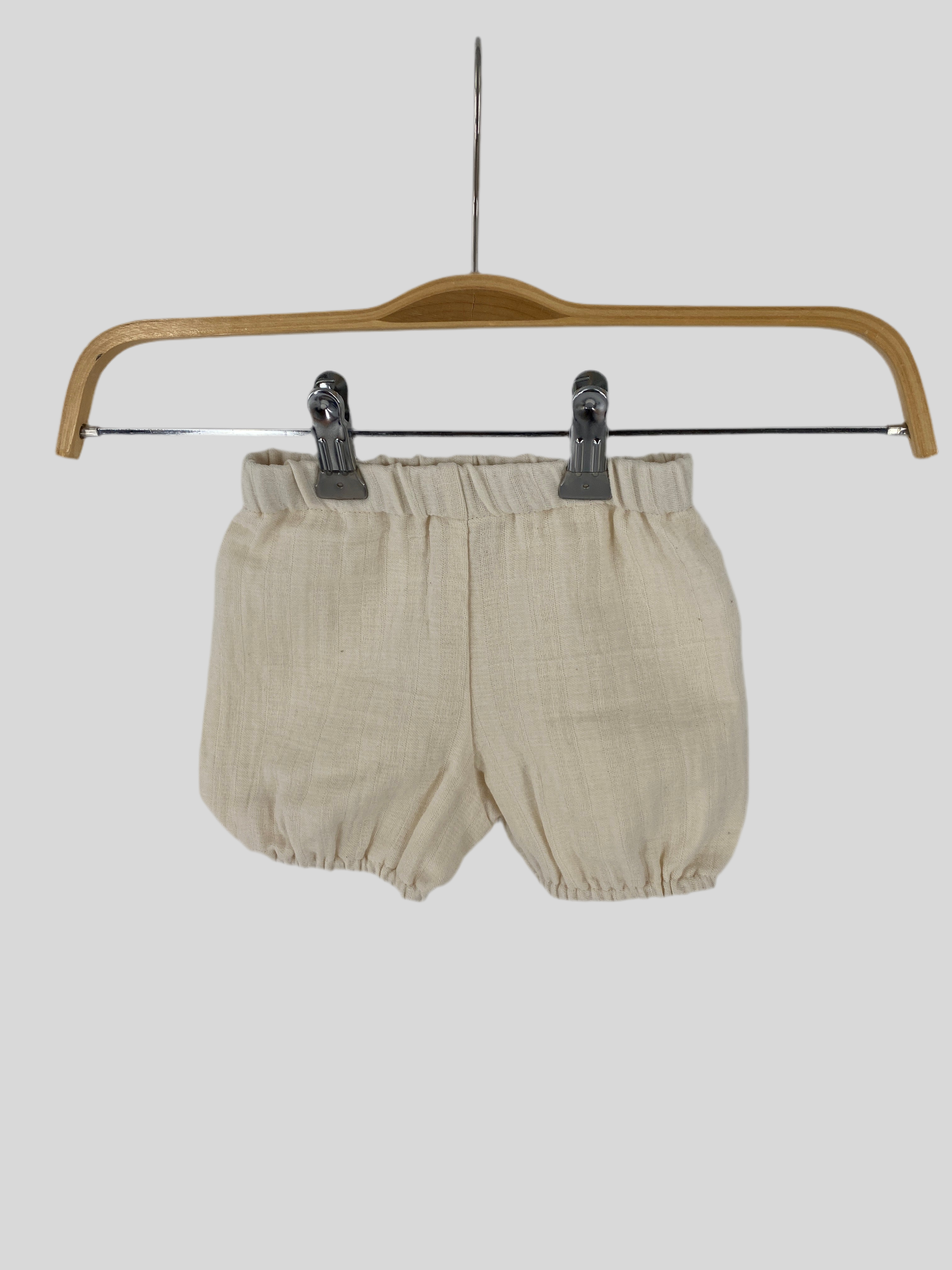 NEU Pumphose Musselin Natur Gr 0M: Weiße Shorts auf einem Kleiderbügel, mit schwarzen Clips befestigt, vor einem metallischen Hintergrund. Ideal für modische, natürliche Baby-Outfits.