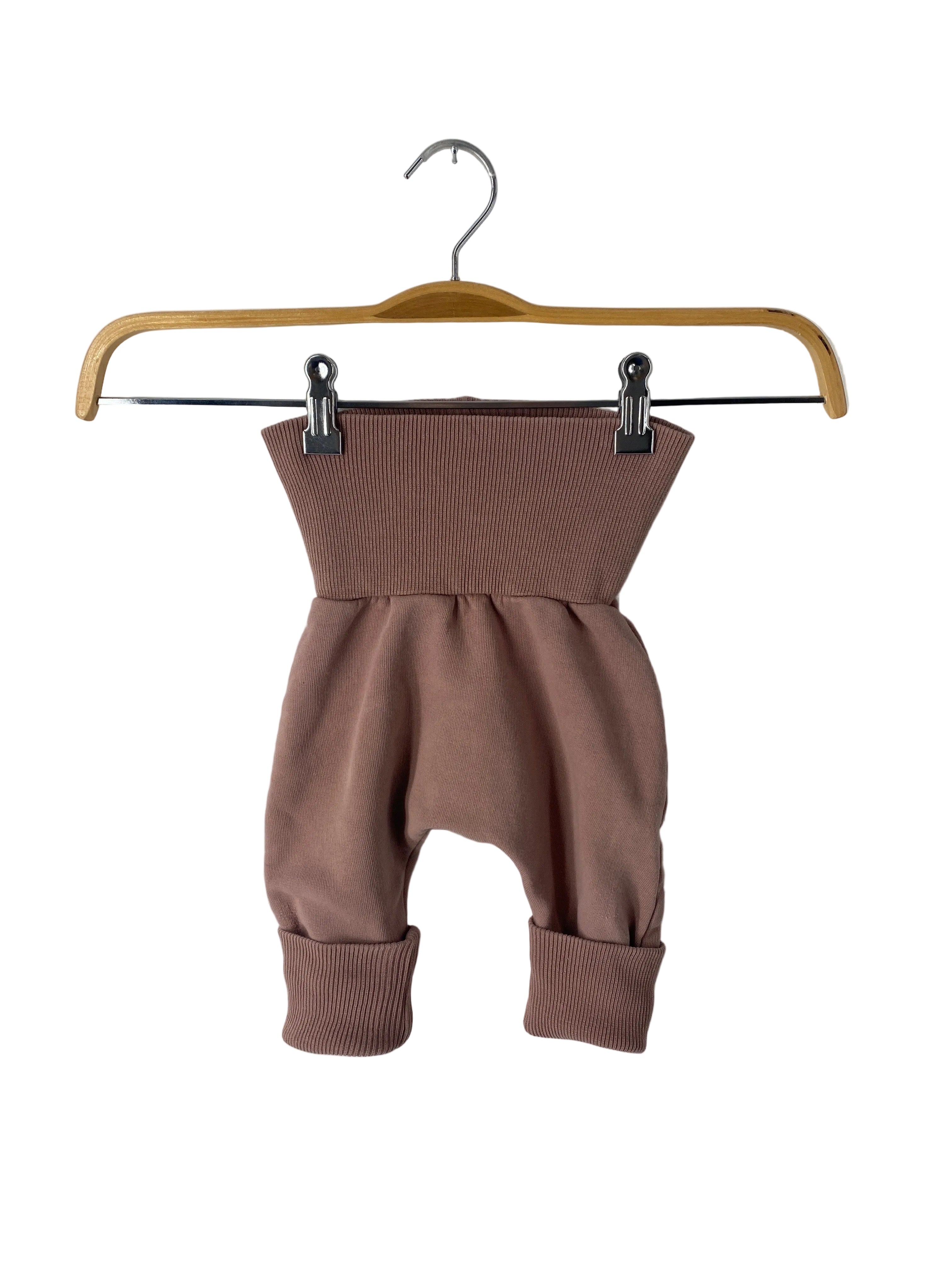Wollwalk Mütze Jade mit Teddyfutter Gr 1Y: Kleine Baby-/Kleinkindhose im Harem-Stil, mit breitem, geripptem Bund und Bündchen, in gedämpftem Rosabraun, an einem Holzkleiderbügel mit Metallklammern hängend.