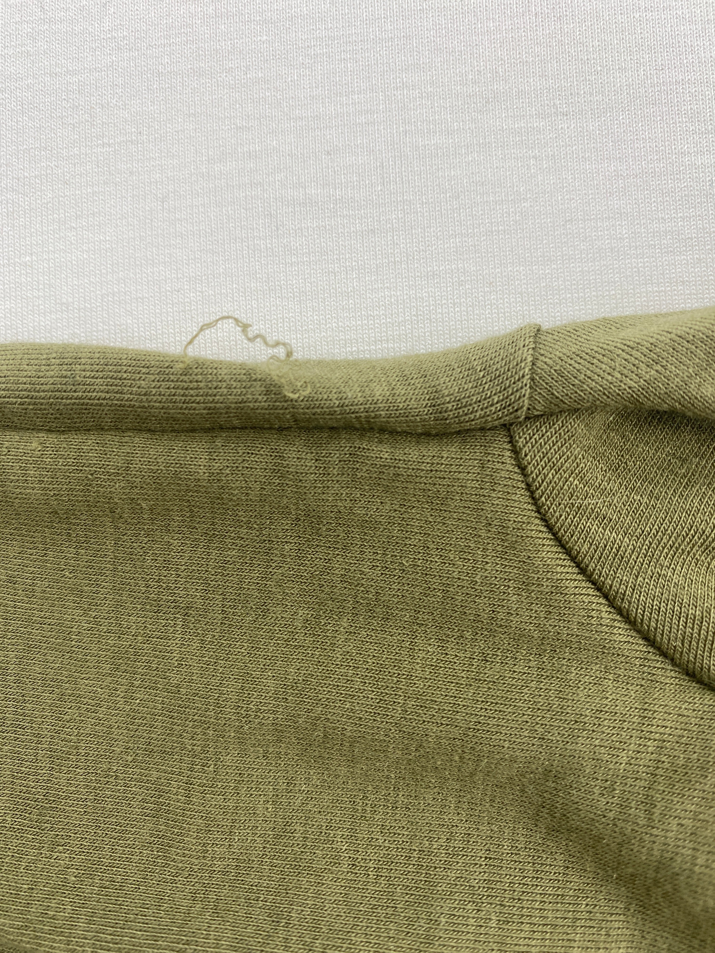 Nahaufnahme des Loopbody Kaschmir Mix Pistazie Gr Mini, zeigt ein leichtes Strickmuster mit einem losen Fadenzieher am Halsausschnitt, auf einem weißen Untergrund liegend. Zustand: neu.