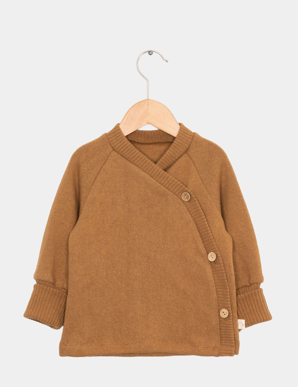 Kopie Baby Schlüttli Strick Sahara Gr 6M: Ein kleiner, langärmeliger Wickelstrickcardigan mit V-Ausschnitt und asymmetrischer Knopfleiste, befestigt mit drei Holzknöpfen, präsentiert auf einem hellen Holzbügel. Zustand: neu.