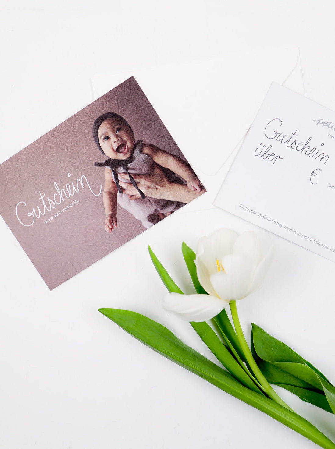 (Kopie) Geschenkgutschein: Zwei Gutscheinkarten, eine mit lächelndem Baby, eine zum Ausfüllen, neben einer weißen Tulpe. Einlösbar online und in Berlin-Tempelhof.
