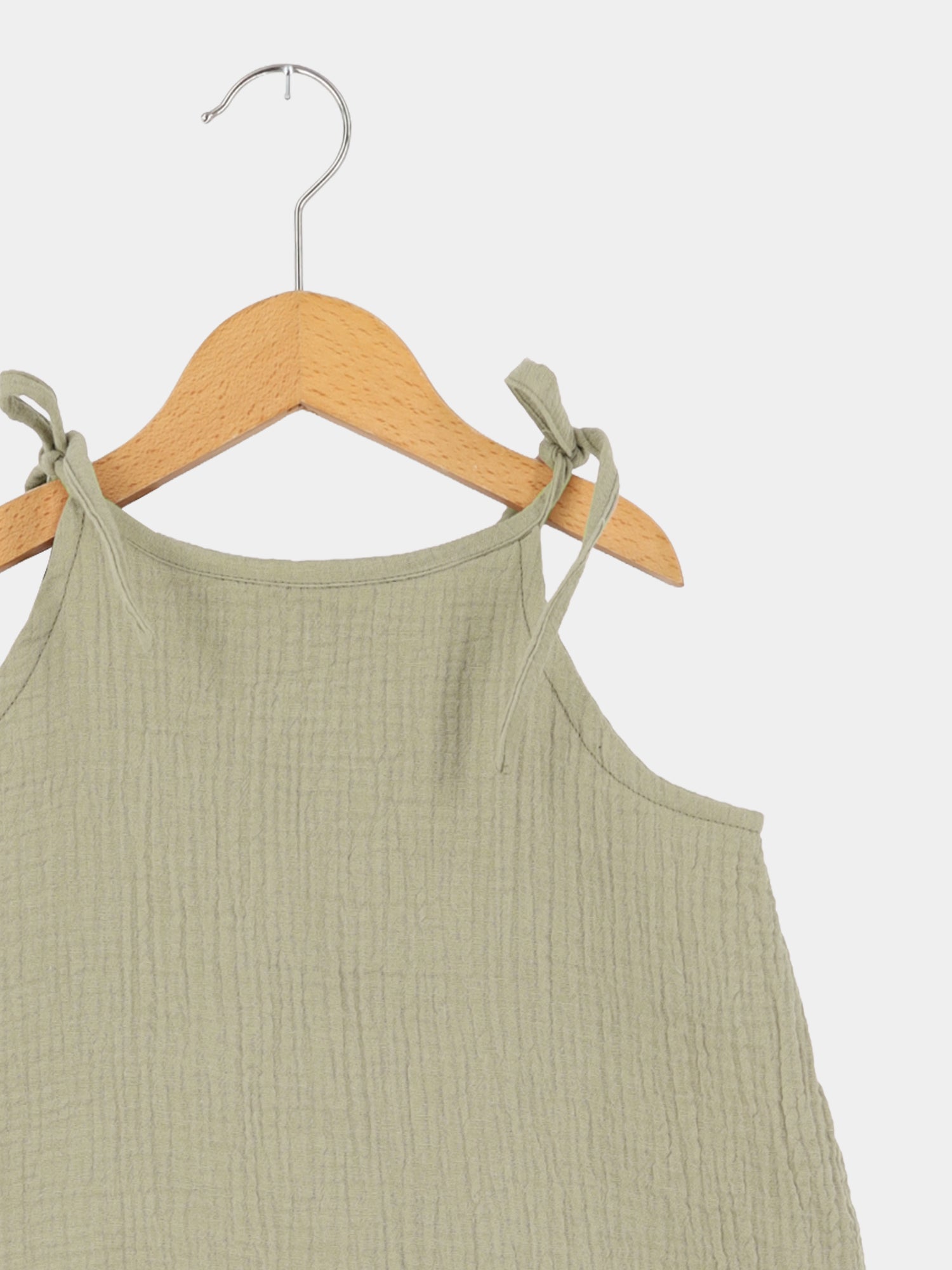 Hängerchen Musselin - Salbei: Ein luftiges, ärmelloses Kleid aus geknitterter Baumwolle mit dünnen, verstellbaren Trägern, die als Schleifen gebunden sind, hängend auf einem Holzkleiderbügel.