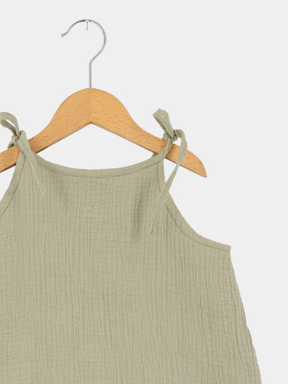 Hängerchen Musselin - Salbei: Ein luftiges, ärmelloses Kleid aus geknitterter Baumwolle mit dünnen, verstellbaren Trägern, die als Schleifen gebunden sind, hängend auf einem Holzkleiderbügel.