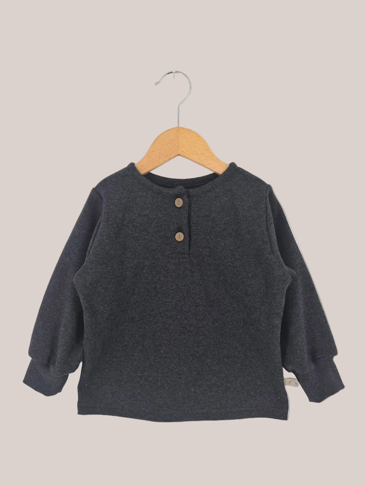 Hemdchen Feinstrick - Ruß: Ein langärmeliges Henley-Shirt für Kinder, aus weichem Feinstrick mit rundem Ausschnitt und zwei Holzknöpfen, auf einem Holzbügel vor weißem Hintergrund.