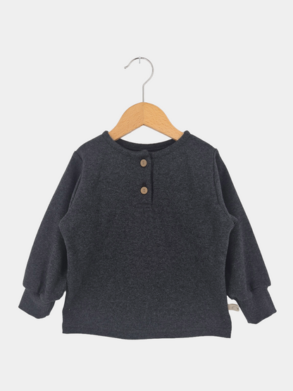 Hemdchen Feinstrick - Ruß: Ein langärmeliges Henley-Shirt für Kinder, aus weichem Feinstrick mit rundem Ausschnitt und zwei Holzknöpfen, auf einem Holzbügel vor weißem Hintergrund.