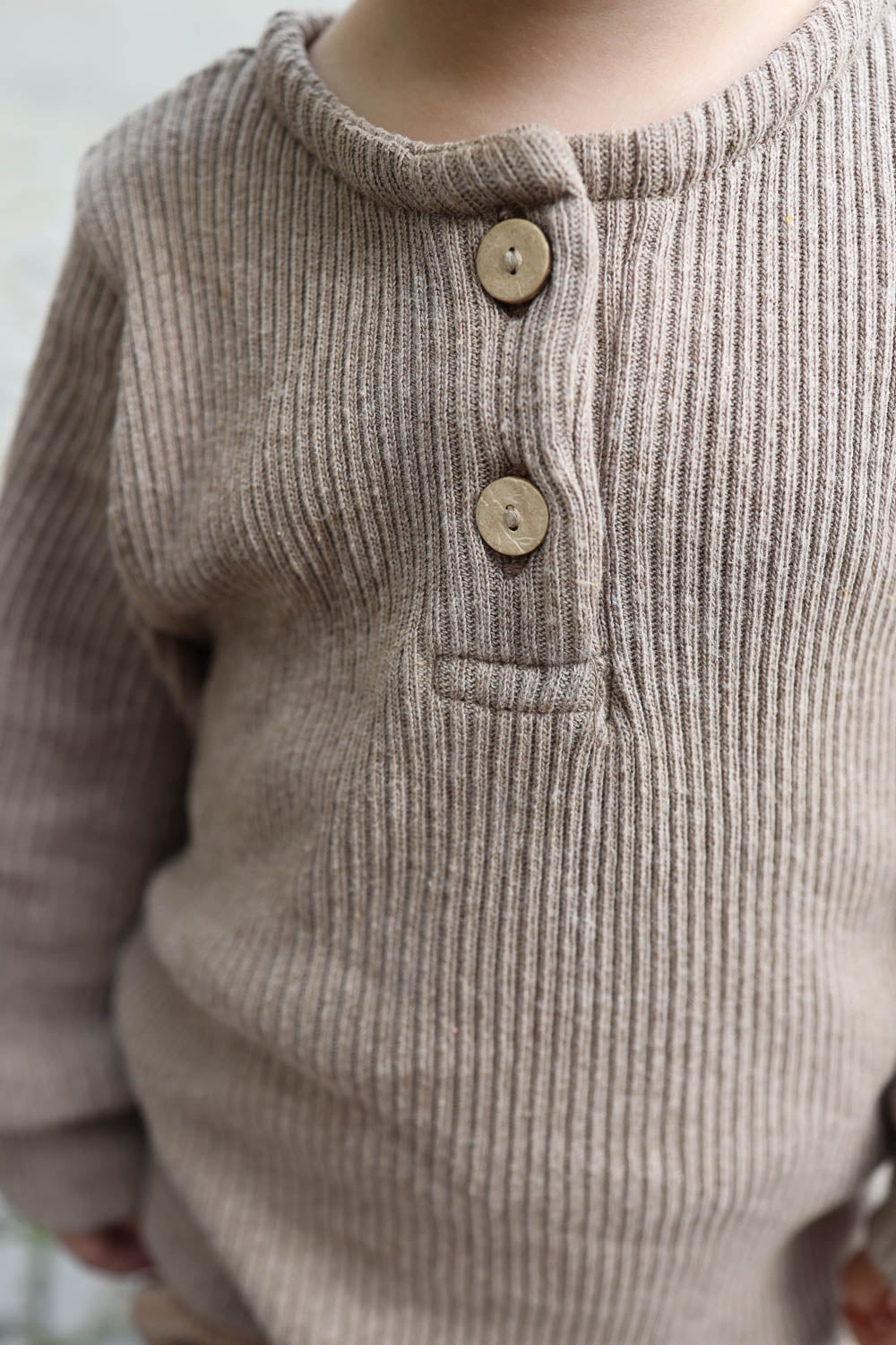 Kind trägt Hemdchen Rippstrick - Hafer, zeigt Nahaufnahme von Knöpfen und geripptem Strickstoff. Unisex, aus recycelter Baumwolle, ideal für Lagenlooks.