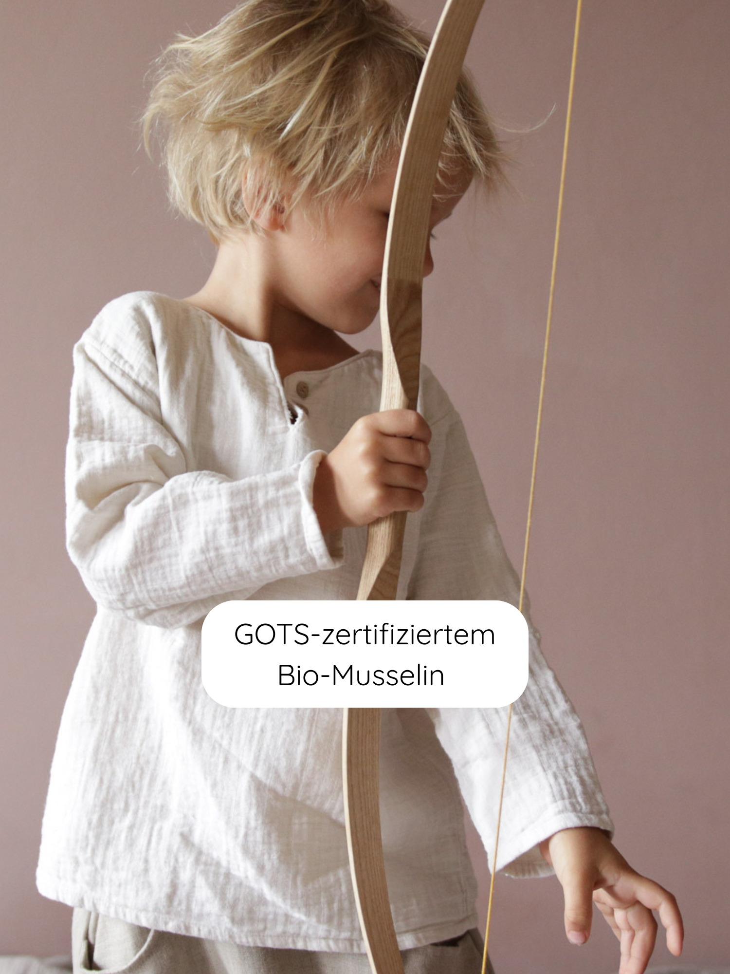 Kleines blondes Kind in lockerem Hemdchen Bio-Musselin - Natur, aus GOTS-zertifiziertem Bio-Musselin, hält einen einfachen Holzbogen. Ideal für warme Tage, bietet Bewegungsfreiheit und Komfort.