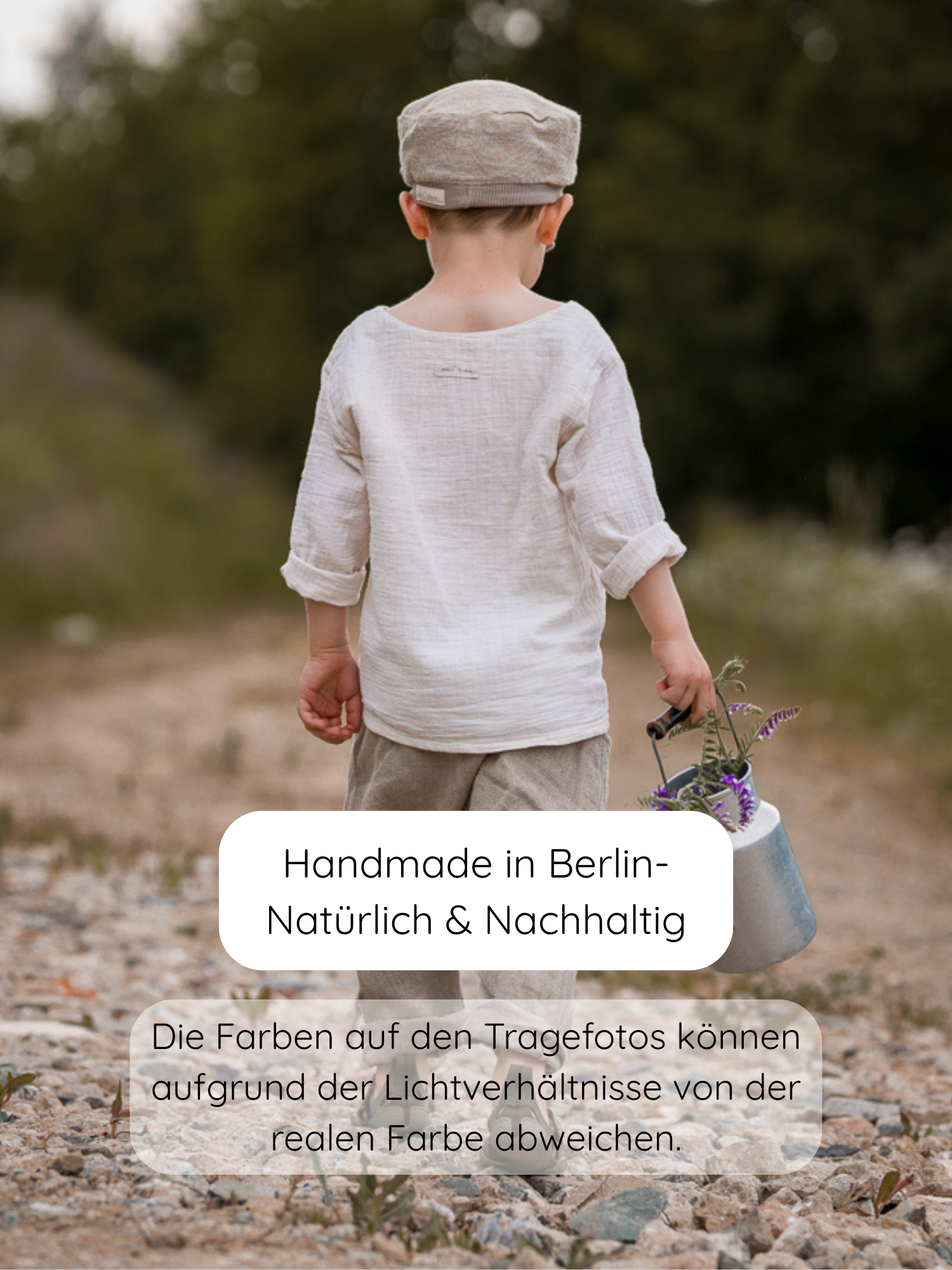 Kleinkind von hinten auf einem Pfad, trägt Hemdchen Bio-Musselin - Natur, mit lockerem Schnitt, hält Metallgießkanne mit lila Wildblumen. Kleidung aus Berlin, handgemacht, nachhaltig.