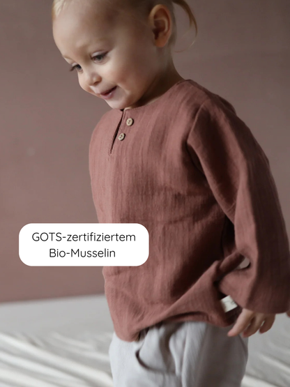 Kleinkind trägt Hemdchen Bio-Musselin - Zimt, ein langärmeliges, rostbraunes Oberteil aus weichem, geknittertem GOTS-zertifiziertem Bio-Musselin, ideal für warme Tage, mit lockerer Passform und atmungsaktiver Qualität.