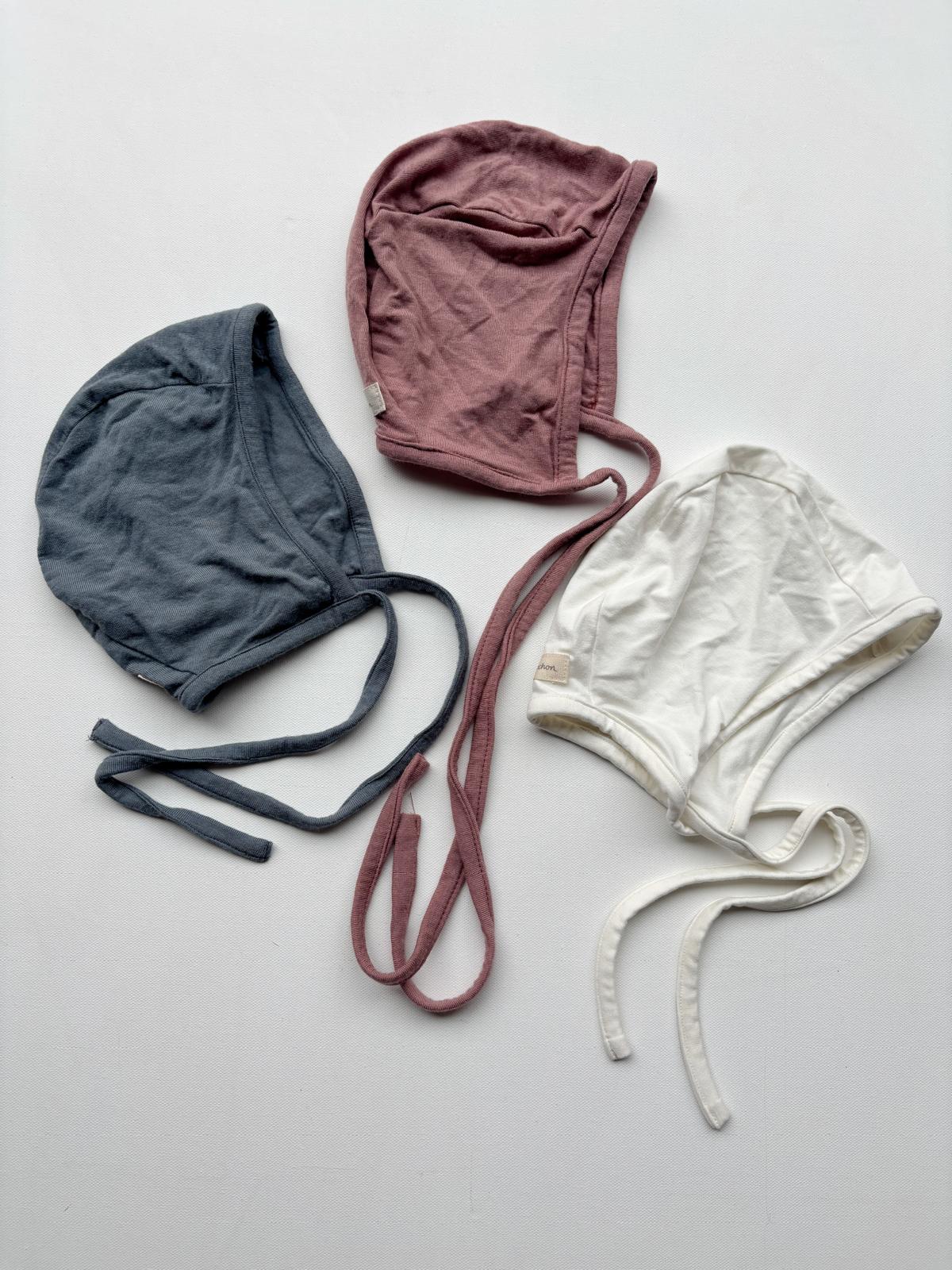 Set 12 drei Häubchen in Gr 2Y, bestehend aus mehreren Babyhäubchen, darunter ein graues mit Bändern und ein weißes mit Schnur, auf einer neutralen Oberfläche präsentiert. Zustand: neu.