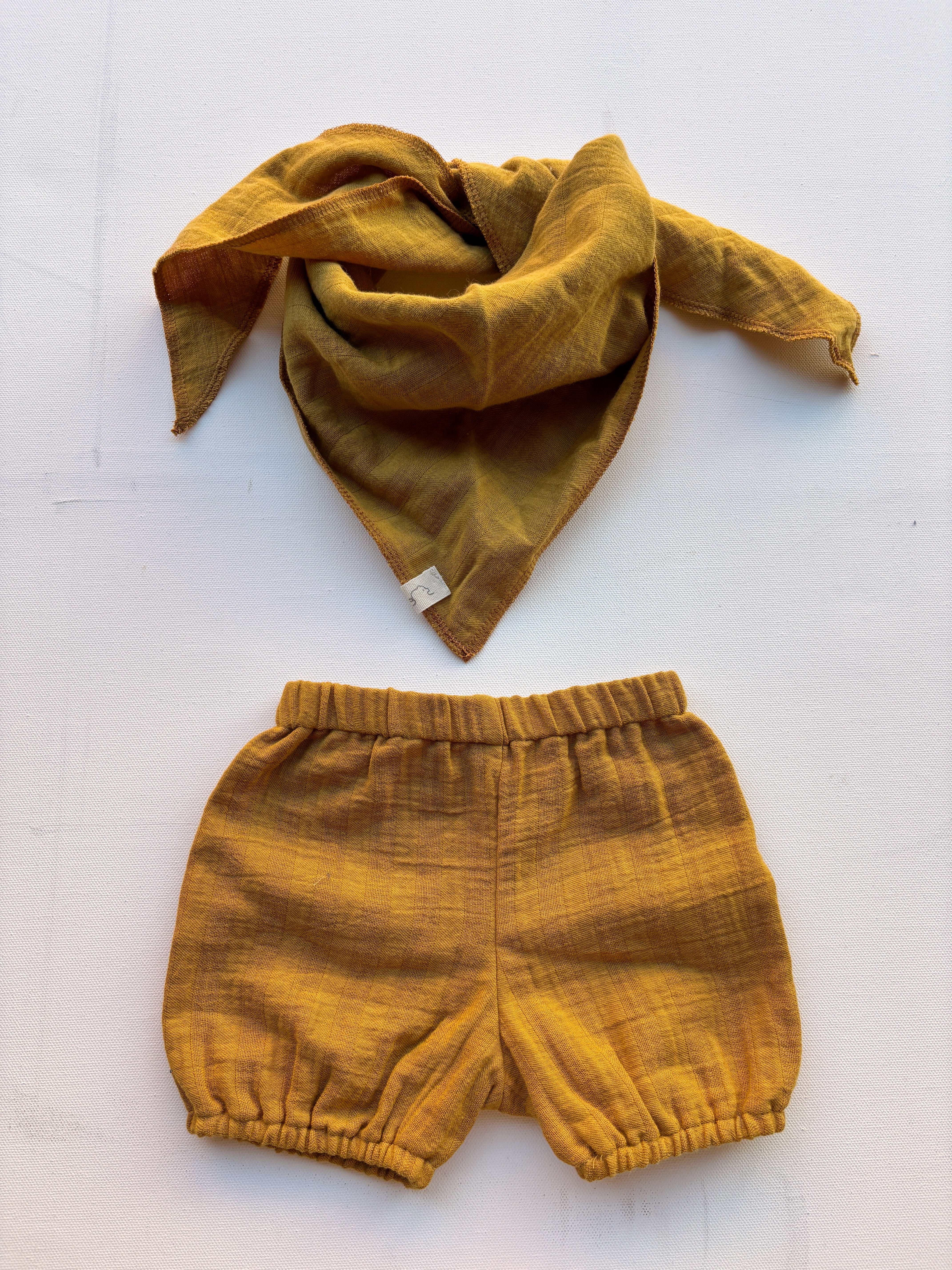 Baby Set Dreieckstuch & Pumphose Bio-Musselin Kurkuma Gr. 0M, bestehend aus einem Dreieckstuch und Pumphose, auf einer weißen Oberfläche präsentiert. Beide Teile sind aus weichem Musselin-Stoff.