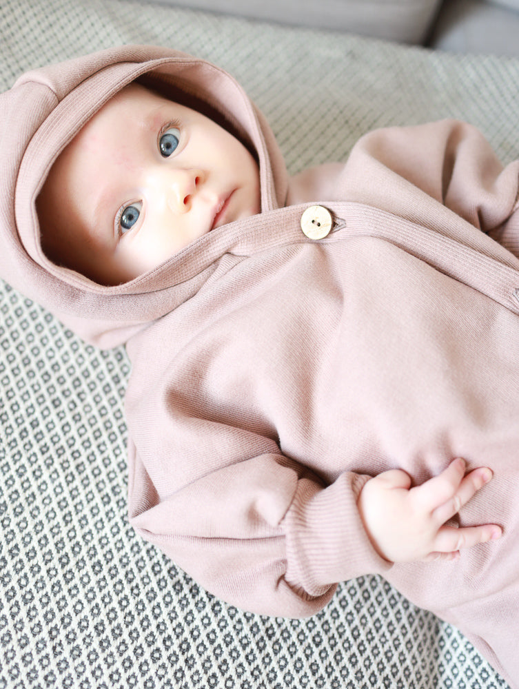 Ein Baby trägt den Kuschelanzug Bio-Sweat - Puder, einen weichen, leicht angerauten Overall mit Wichtelkapuze, ideal für Komfort und Wärme, während es entspannt auf einer gemusterten Decke liegt.