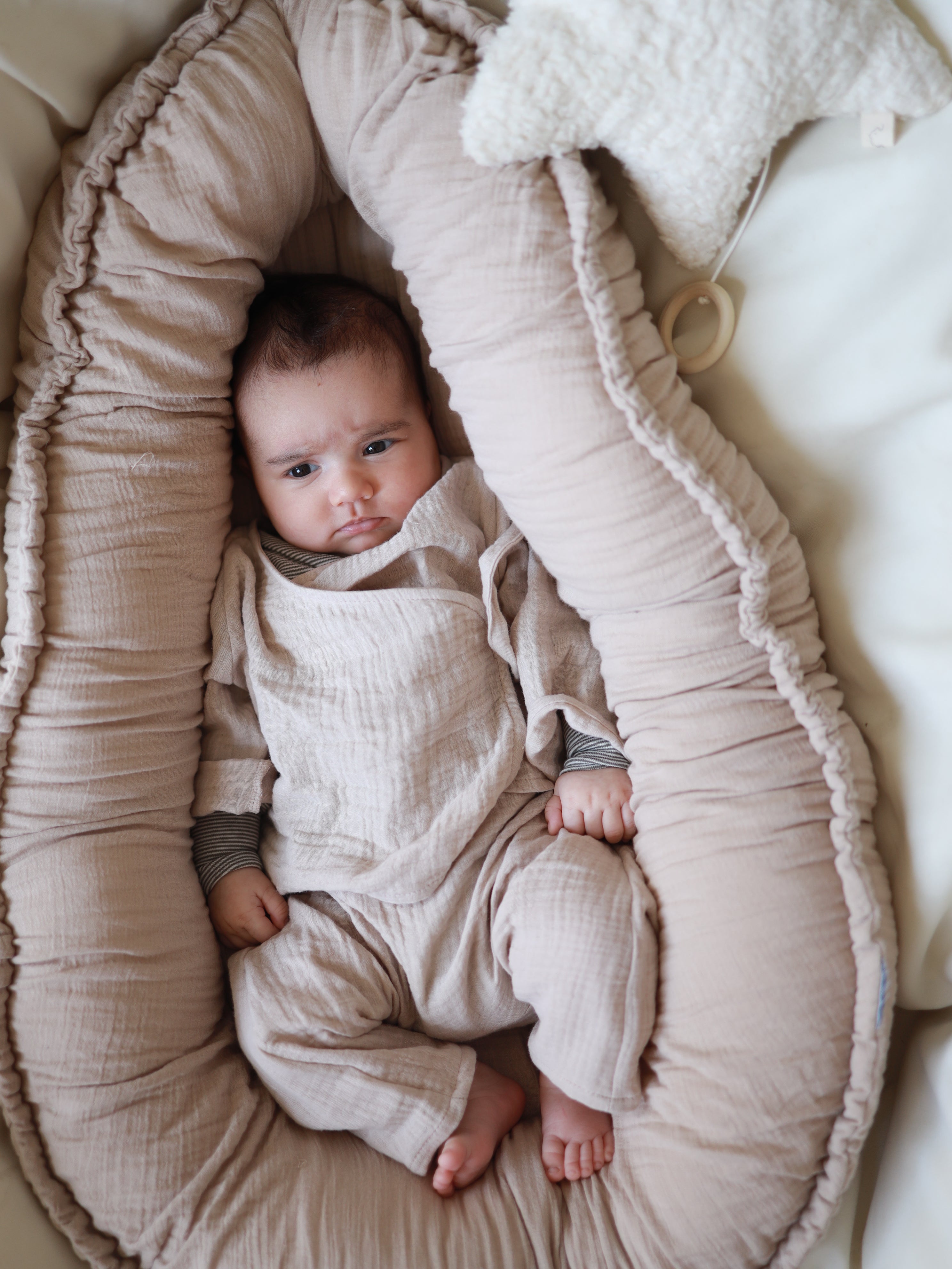 Baby-Set Baumwoll-Musselin - Sand: Ein Baby liegt in einem weichen, gepolsterten Lounger, trägt ein Wickel-Schlüttli und Hose aus atmungsaktivem Musselin. Ein sternförmiges Plüschkissen hängt oben rechts.
