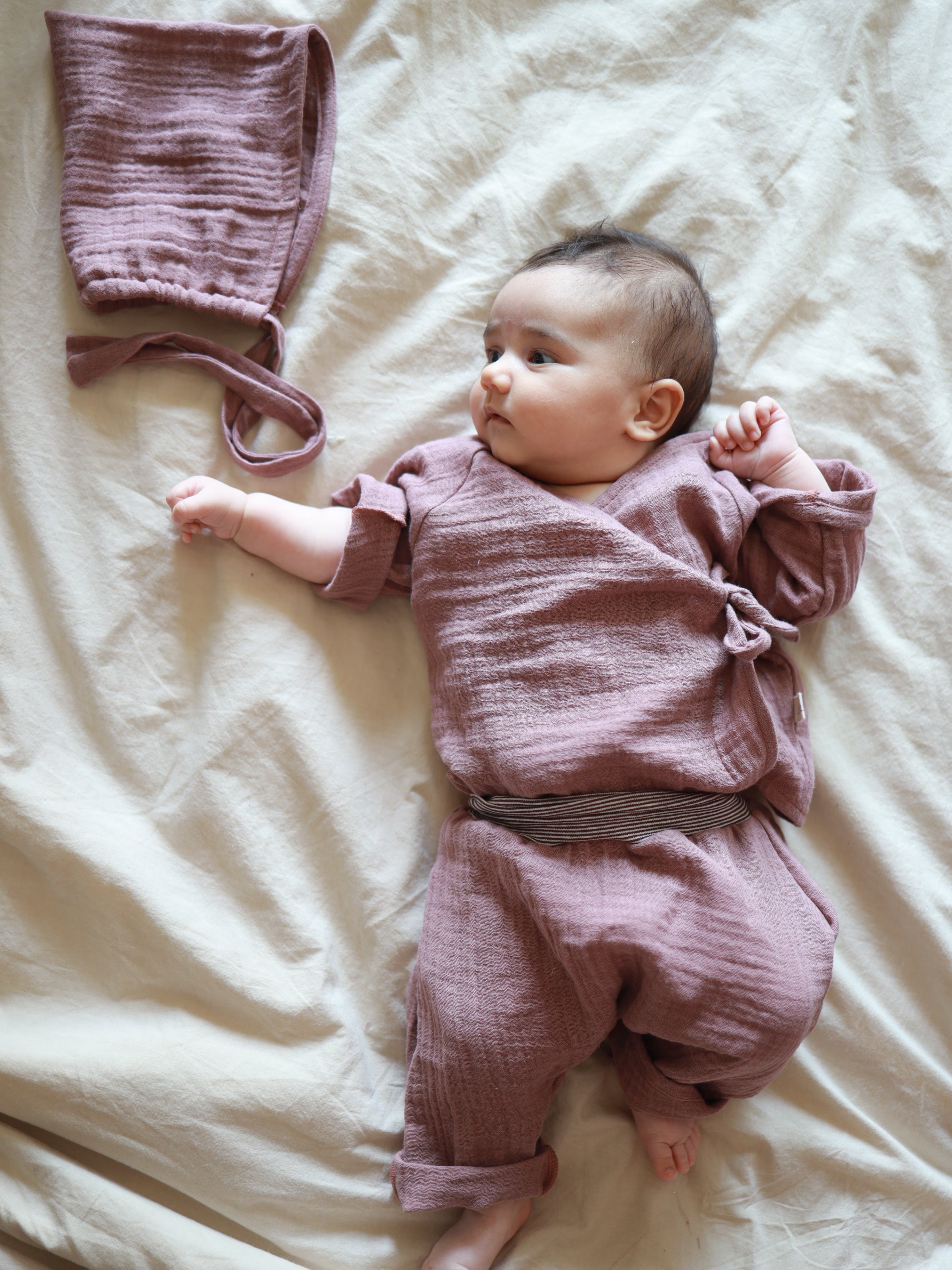 Baby-Set Baumwoll-Musselin - Malve: Ein Baby liegt auf einem hellen Tuch, trägt ein Wickel-Schlüttli mit passender Hose und daneben liegt ein passender Hut aus weichem Musselin-Stoff.