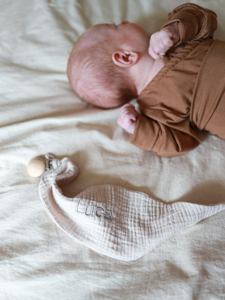 Neugeborenes in braunem Strampler neben einem muslin Schnullertuch, bestickt mit Luca. Individuelle Stickerei bietet personalisierte, handgemachte Geschenke, perfekt für besondere Anlässe.