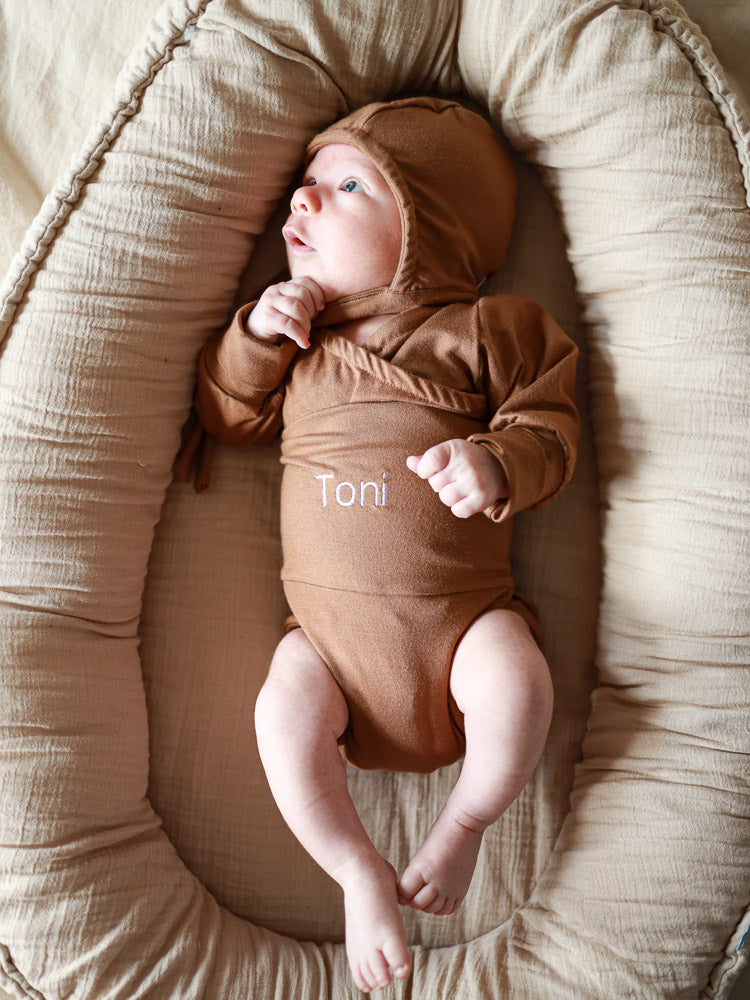 Neugeborenes in braunem Wickel-Body mit „Toni“-Stickerei, liegend auf beigem Nestkissen. Individuelle Stickerei macht es zu einem einzigartigen, personalisierten Geschenk.