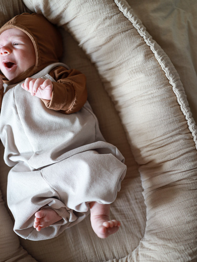 Ein Baby liegt in einem weichen Nest und trägt die Kurze Baby Schlupflatzhose Feincord - Sand, eine bequeme, leicht anzuziehende Latzhose mit Baumwollripp-Halsbündchen, passend für Babys und Kleinkinder.