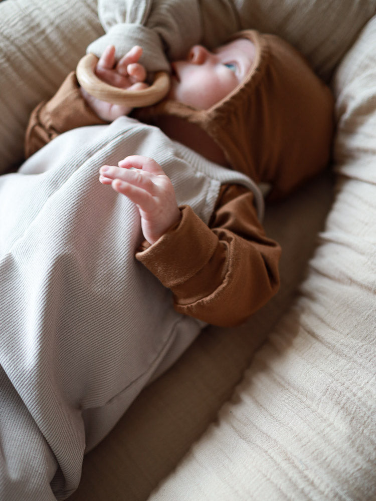 Baby trägt Kurze Baby Schlupflatzhose Feincord - Sand, bequem über braunem Kapuzenoberteil. Es hält ein Holzspielzeug, während es auf einem weichen Kissen liegt. Perfekt für einfaches Anziehen.