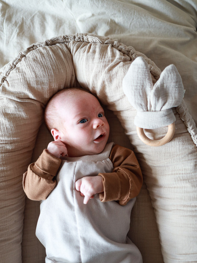 Neugeborenes in einem neutralen Baby-Lounger, trägt eine Kurze Baby Schlupflatzhose Feincord - Sand, daneben ein hölzerner Beißring mit Stoffohren, betont die bequeme und praktische Passform.