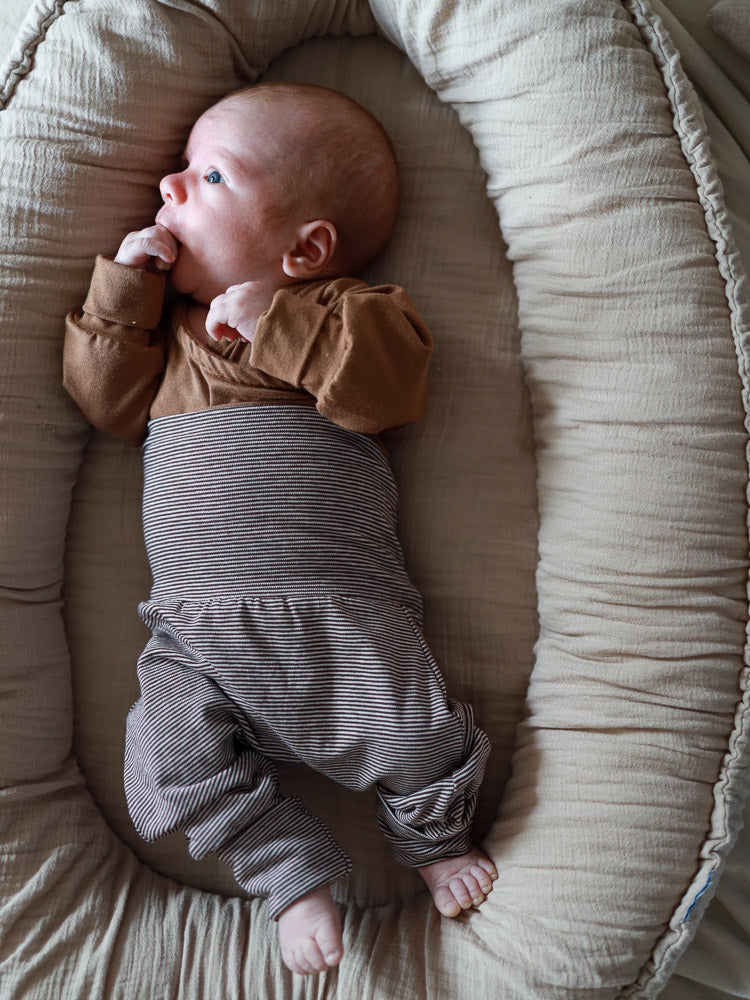 Baby trägt Babyknicker Bio-Baumwolle - Schoko Ringel, liegt im beigen Babynest. Die weiche, braun-gestreifte Hose bietet Bewegungsfreiheit und Komfort mit elastischen Bündchen, ideal für empfindliche Haut.